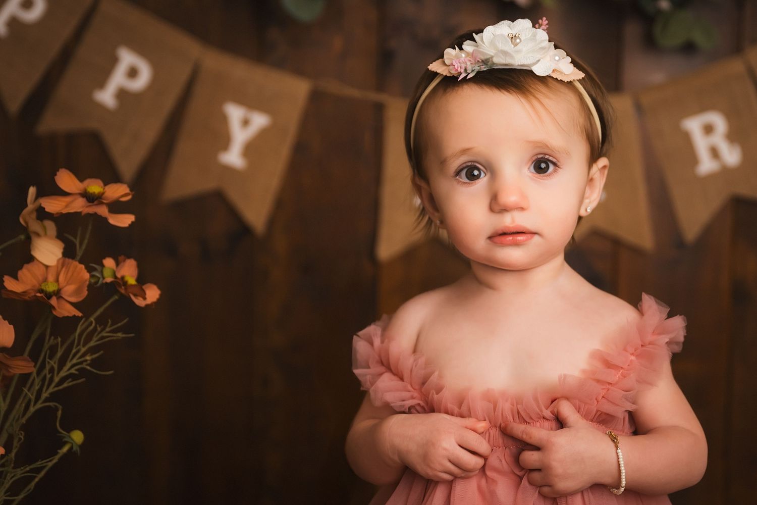 Adorable baby girl in pink tulle dress and floral headband poses for first birthday portrait with rustic wood backdrop.