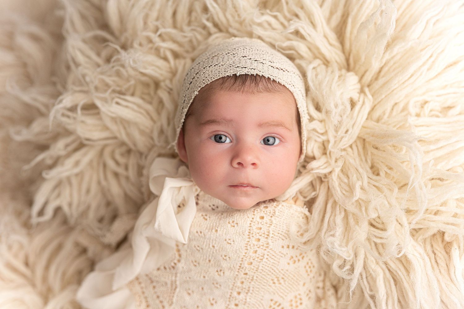 Newborn baby wrapped in cream knit swaddle and bonnet, lying on fluffy white fur backdrop.