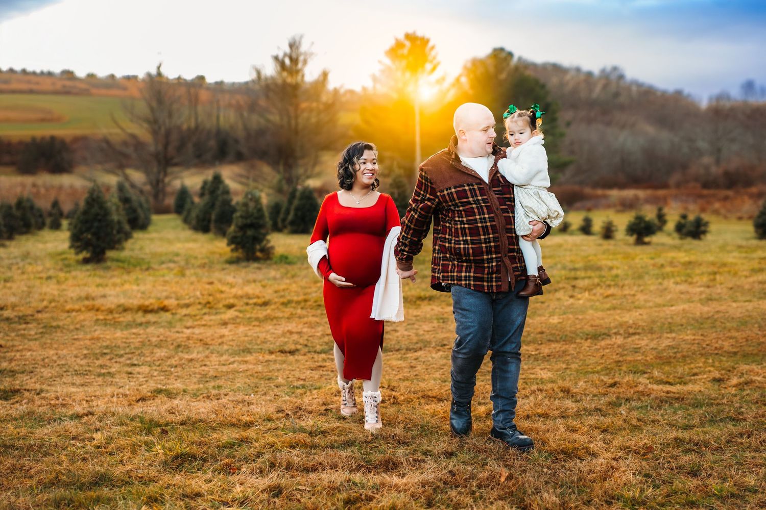 Pregnant woman in red dress with husband and toddler at Christmas tree farm during golden sunset.
