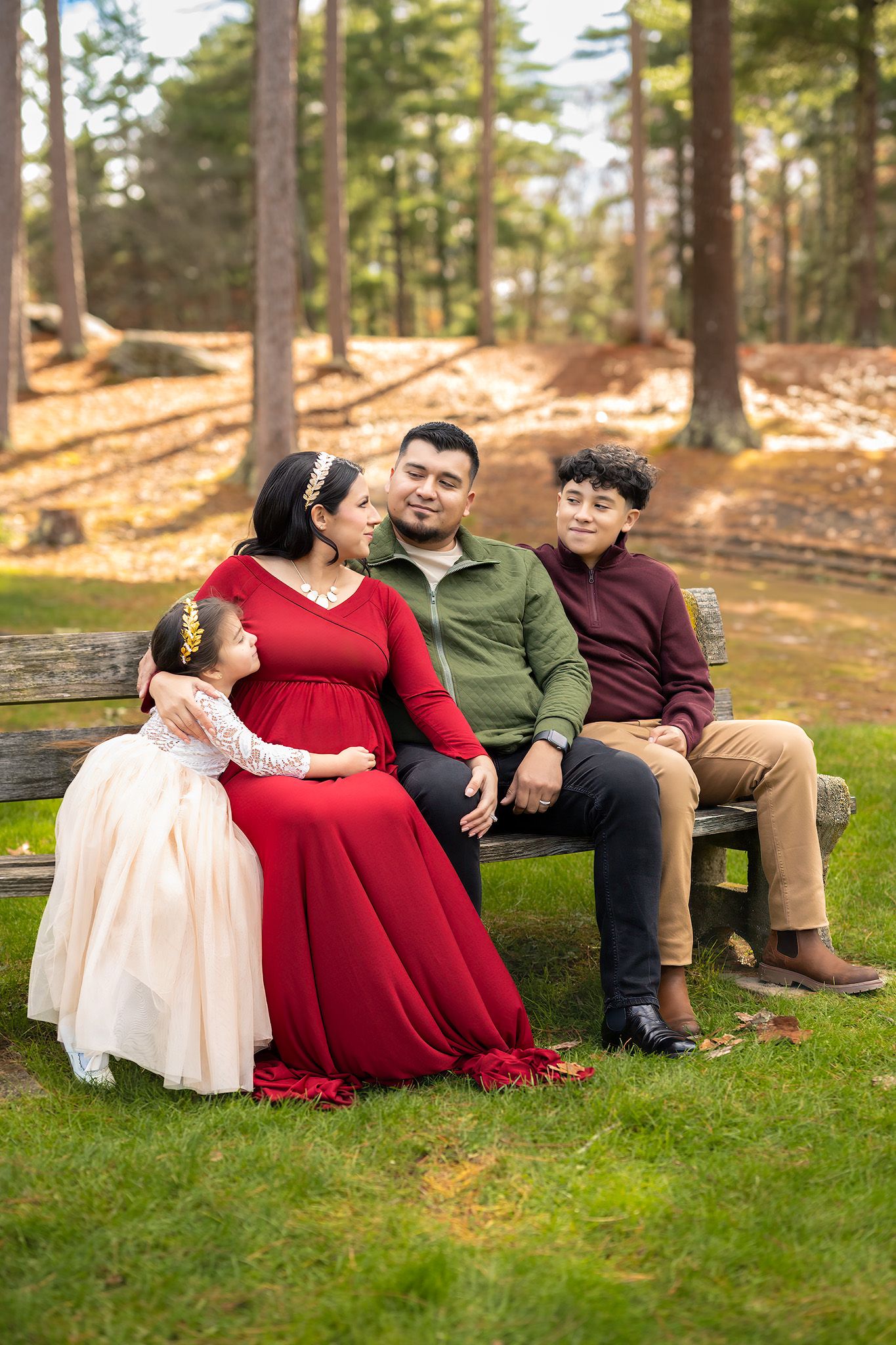 A family of four poses on a bench in a forest park, with a pregnant woman in a red dress.