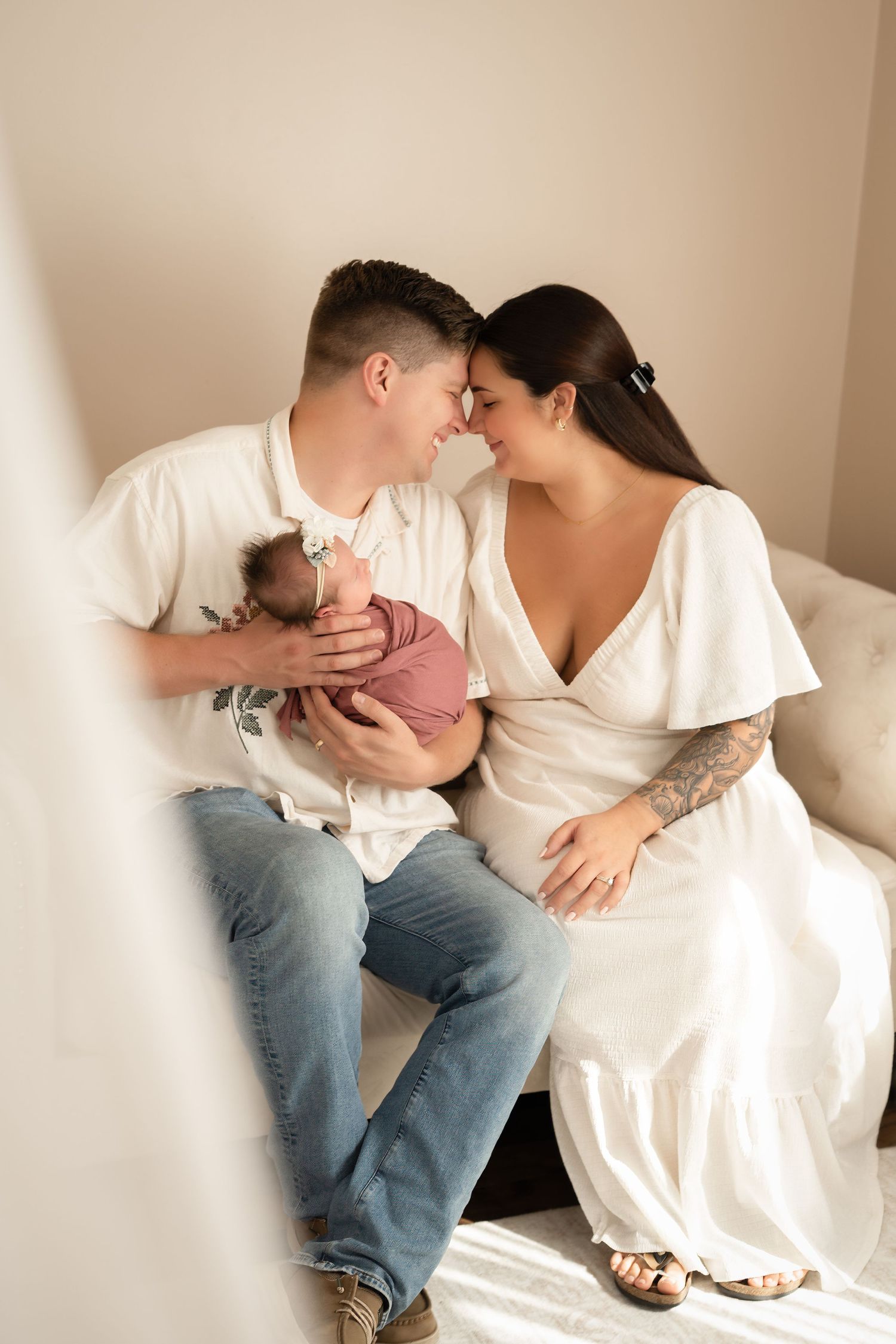 A couple sitting on a bed, touching foreheads, while the man holds their newborn baby wrapped in pink.