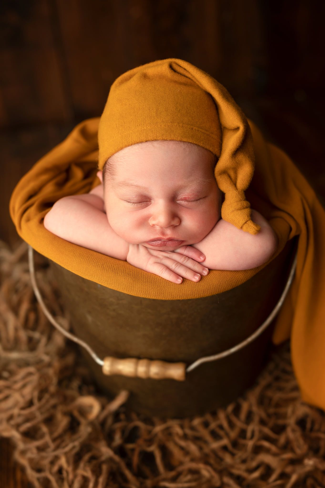 Newborn baby sleeping peacefully in a dark bucket, wearing a mustard yellow knit hat and wrap.