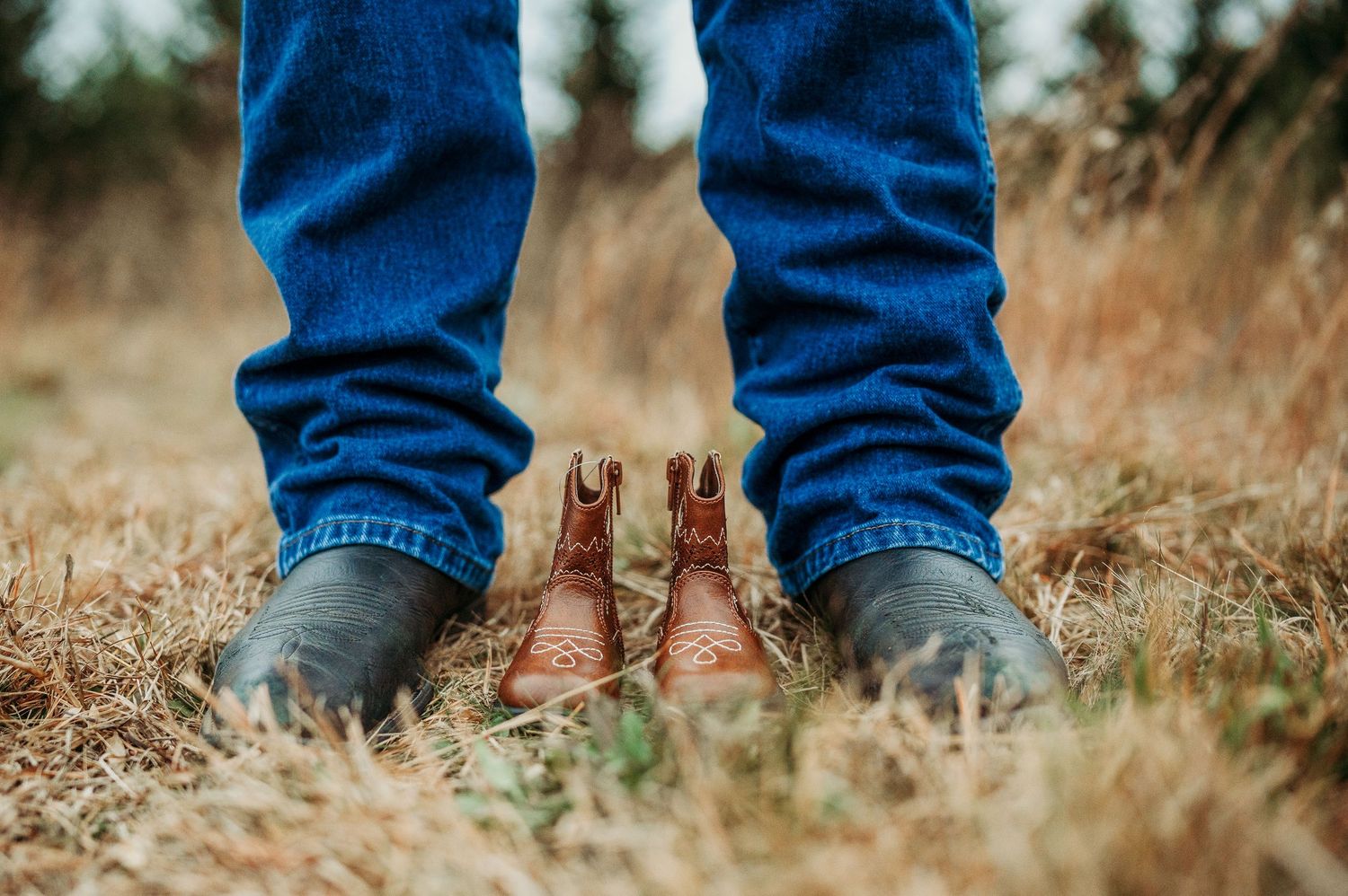Close-up of legs in blue jeans and brown cowboy boots standing on dry grass outdoors.