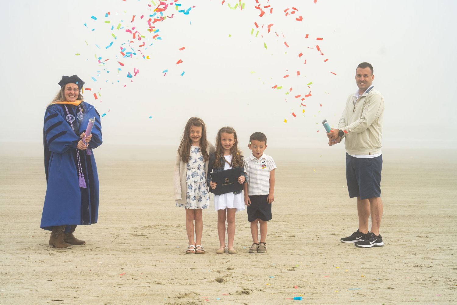 Family celebrates graduation on foggy beach with colorful confetti, graduate in blue cap and gown stands proudly.
