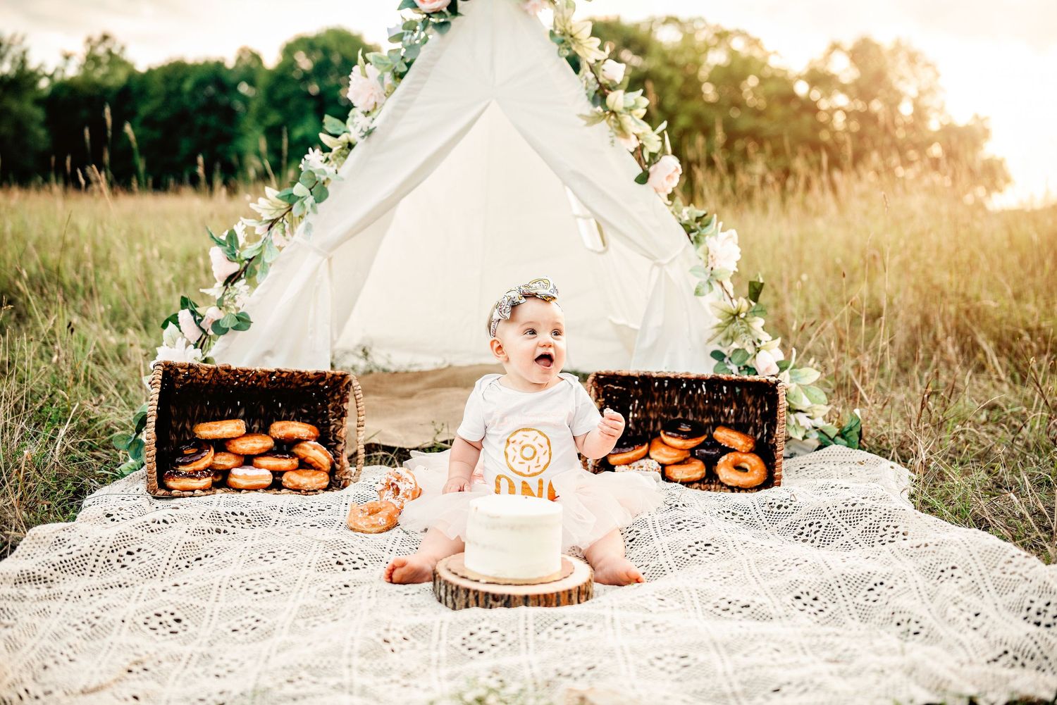 Baby's first birthday cake smash outdoors with donuts, boho teepee, and floral decorations on a lace blanket in a field.