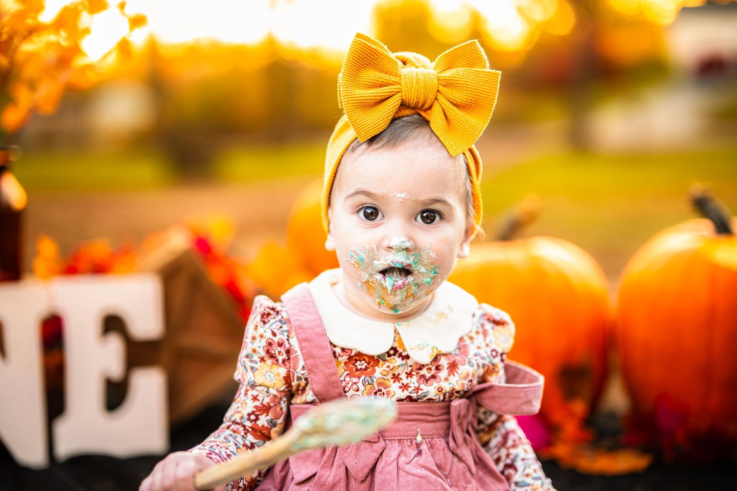 Baby girl in floral dress and yellow bow headband eating cake surrounded by pumpkins in fall outdoor photo session.