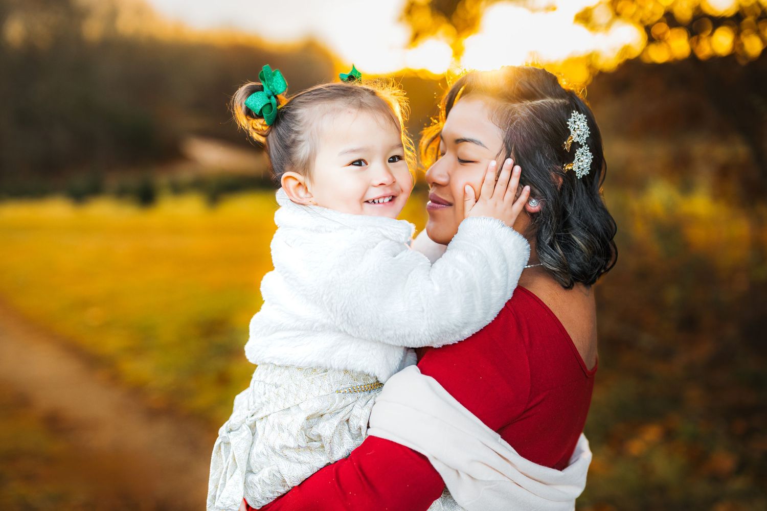 A mother holds her smiling toddler daughter outdoors in warm autumn light, both wearing festive red and white outfits.