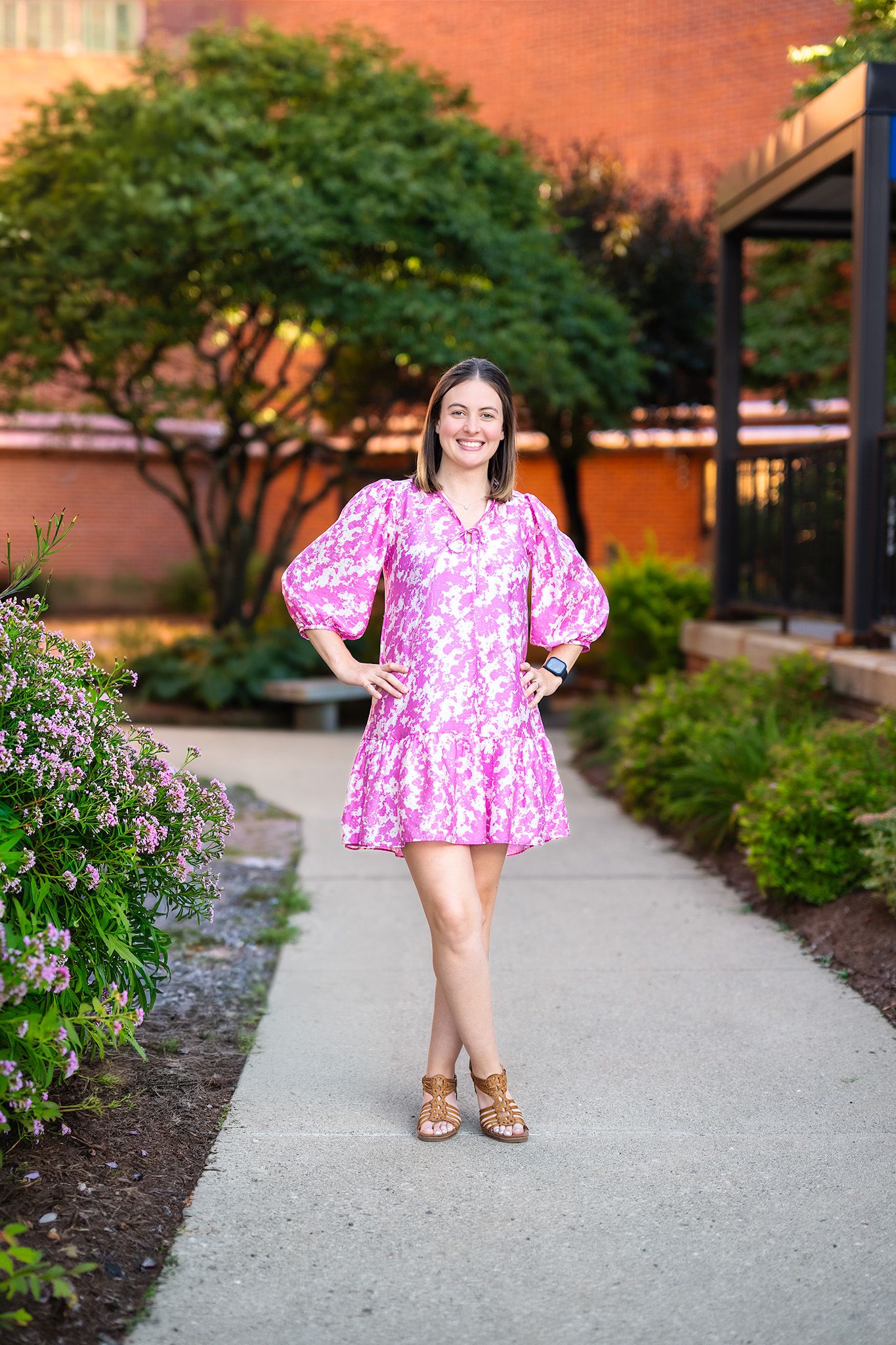Woman in pink floral mini dress with puff sleeves and tan sandals posing on garden path near brick building.