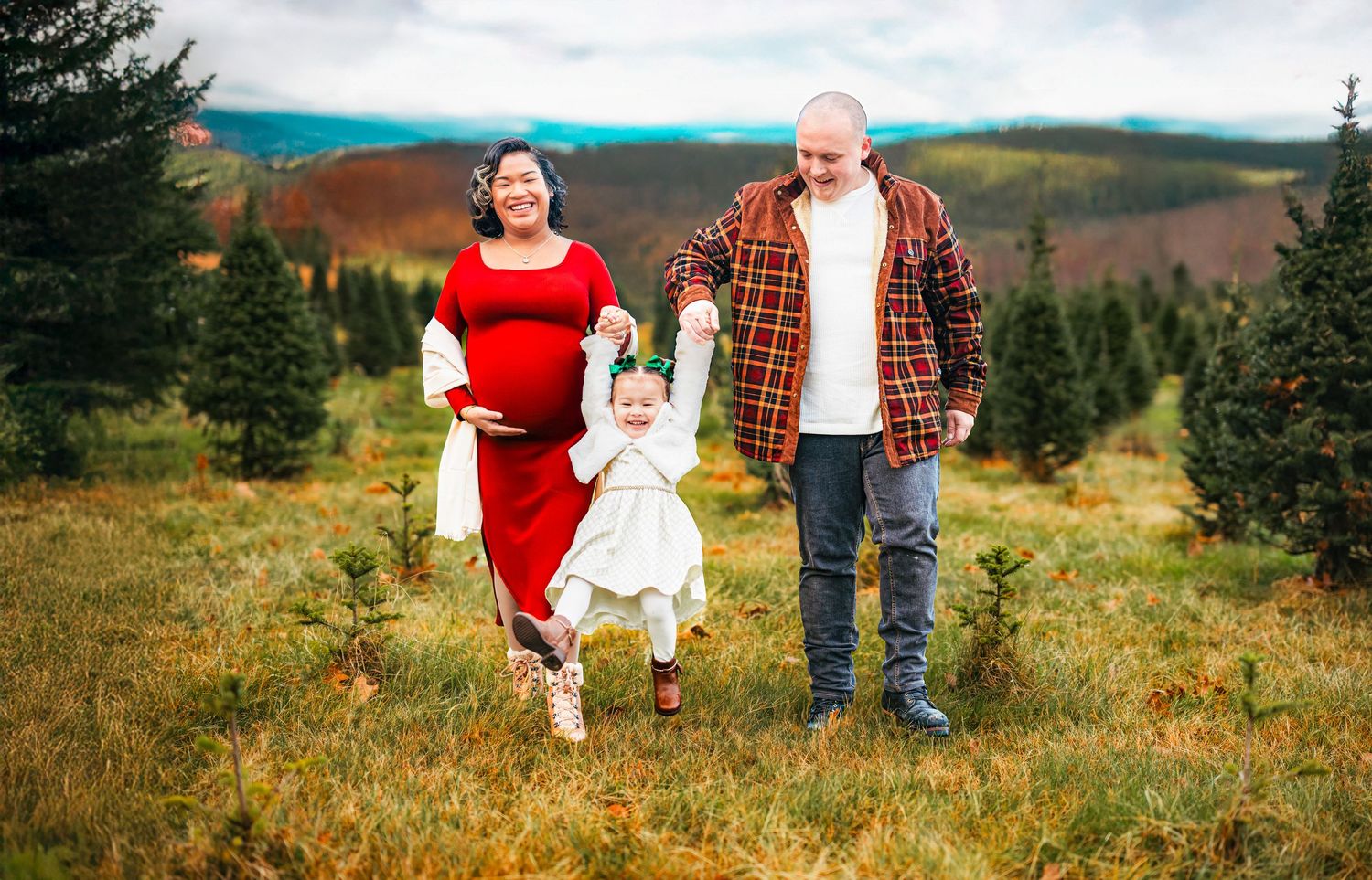 Pregnant woman in red dress with family at Christmas tree farm, man in plaid shirt, toddler in white dress.