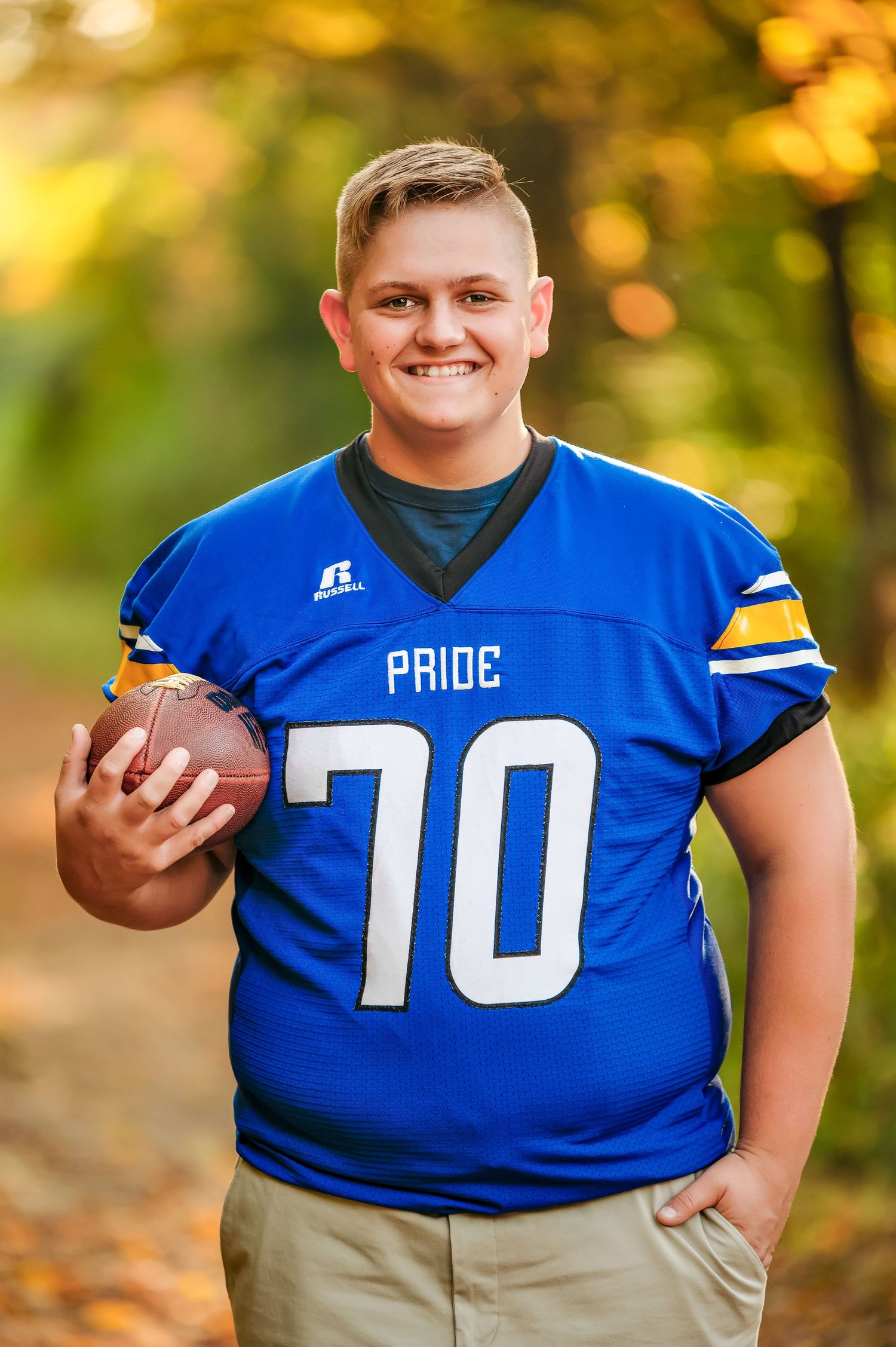 Smiling football player in blue Russell Pride jersey number 70 holding a football outdoors in autumn setting.