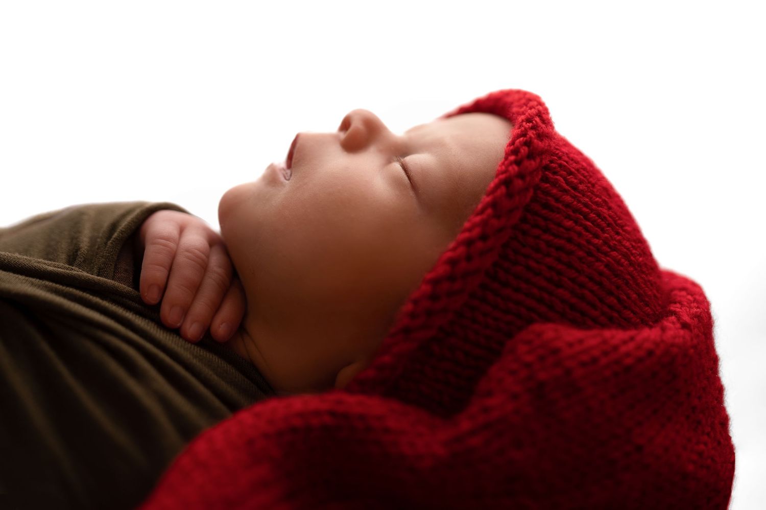 Peaceful newborn baby sleeping wrapped in a red knit blanket and hat, photographed in soft white light.