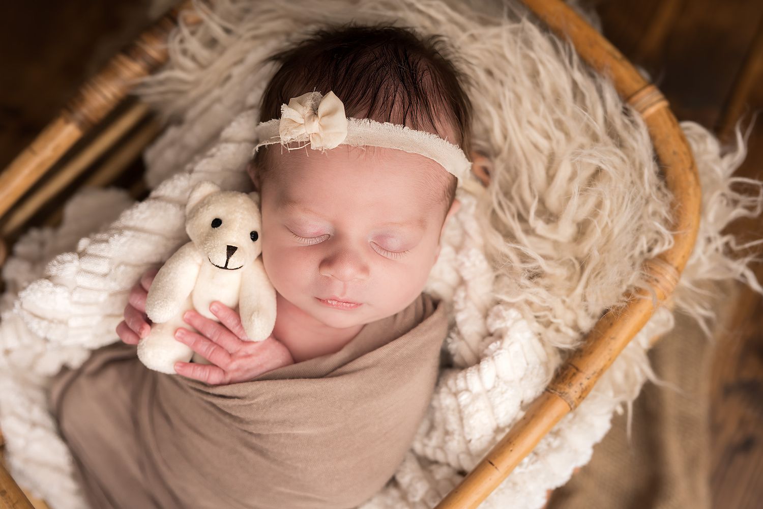Sleeping newborn baby wrapped in cream swaddle holding tiny teddy bear in wicker basket with fur blanket.