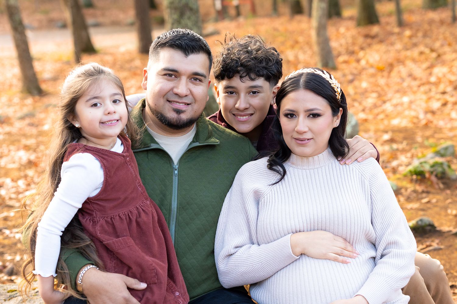 Family of four posing outdoors in autumn forest, pregnant mother holding belly with husband, son, and daughter smiling.