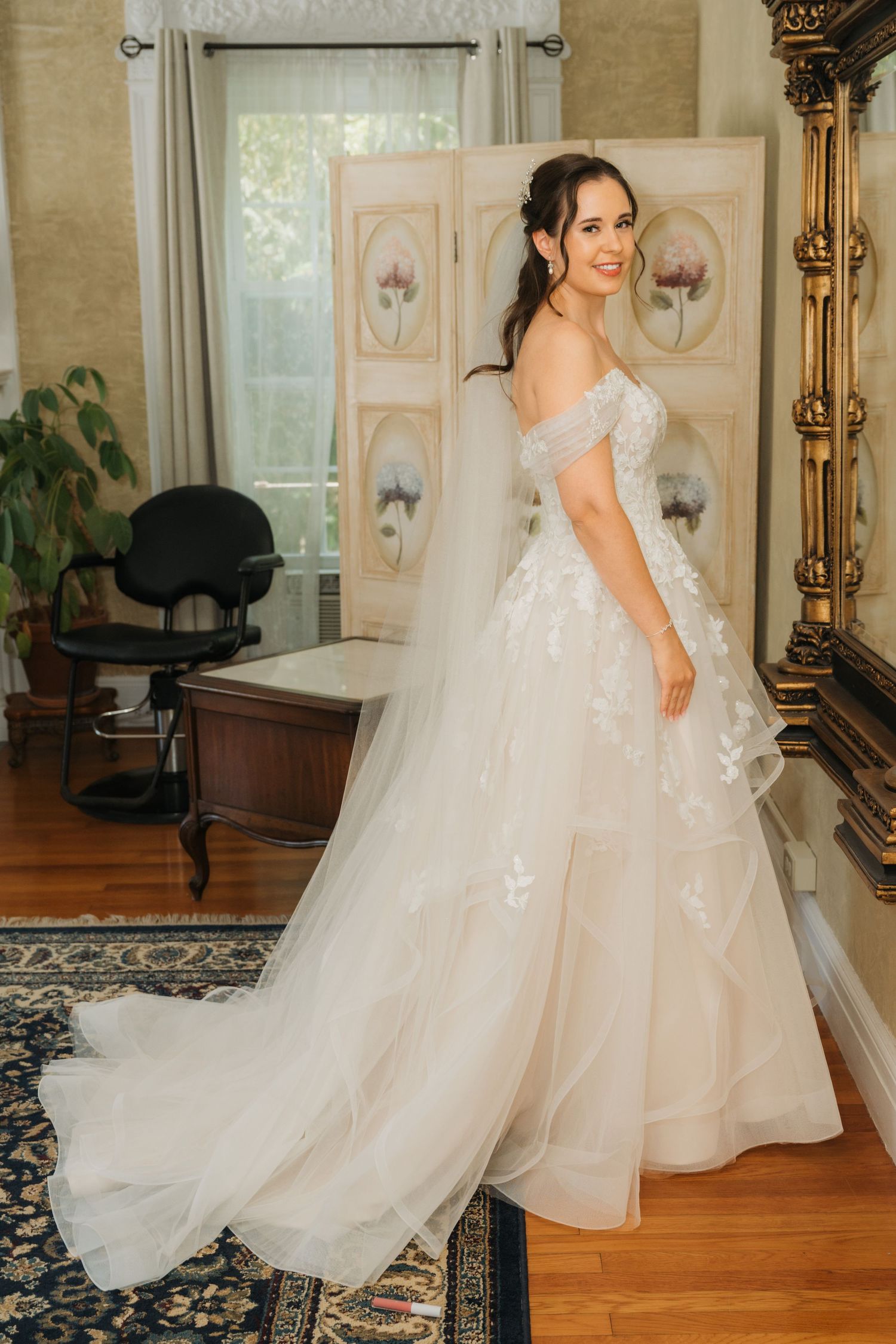 Bride in off-shoulder layered tulle ball gown with floral lace details, posing in elegant room with veil.
