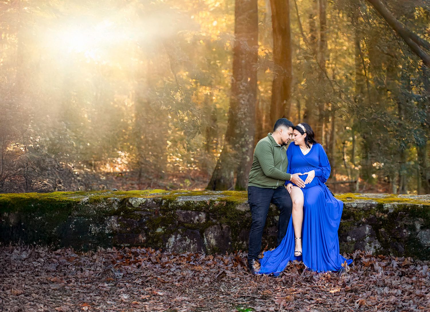 Couple sharing intimate moment outdoors in forest, woman in flowing blue maternity gown, golden sunlight streaming through autumn trees.