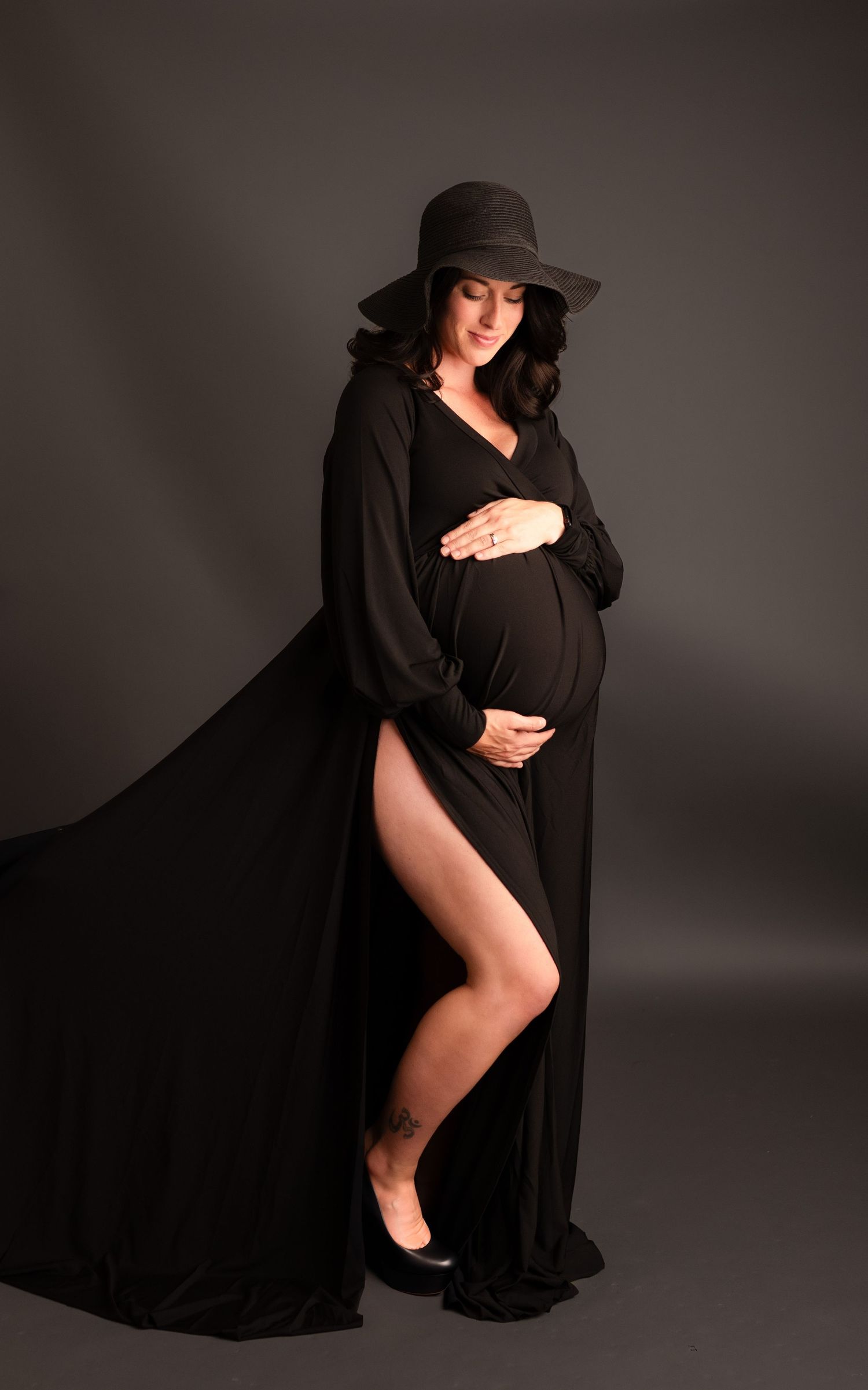 Pregnant woman in elegant black flowing gown and wide-brim hat cradling baby bump in professional studio photo.