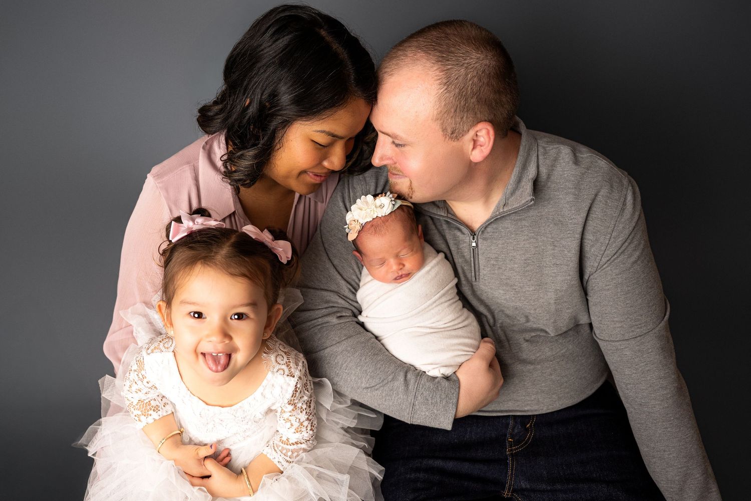Happy family of four with parents and two young children smile together against a dark gray studio backdrop.