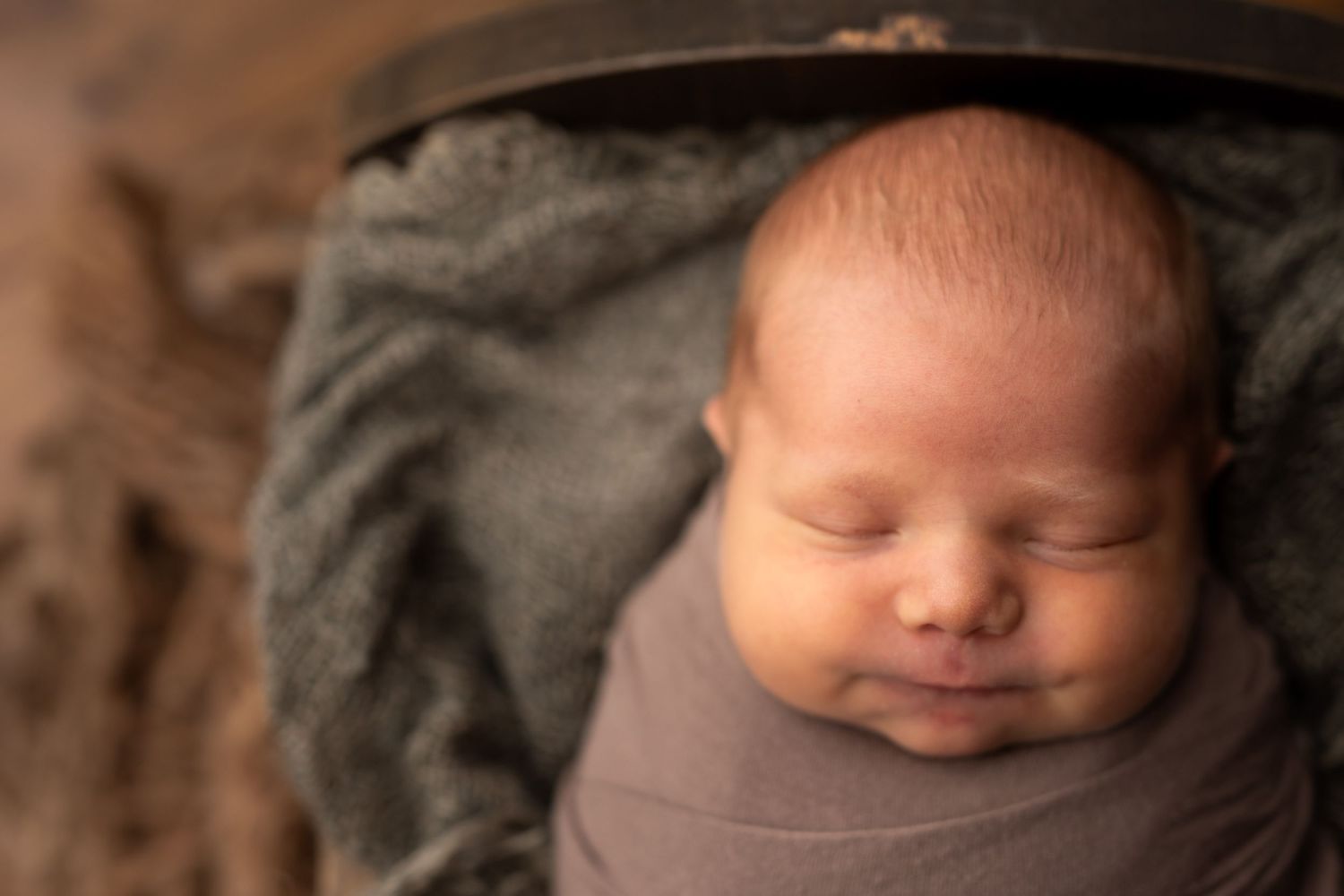 Peaceful newborn baby swaddled in gray wrap, sleeping in a rustic wooden bowl with soft green blanket.