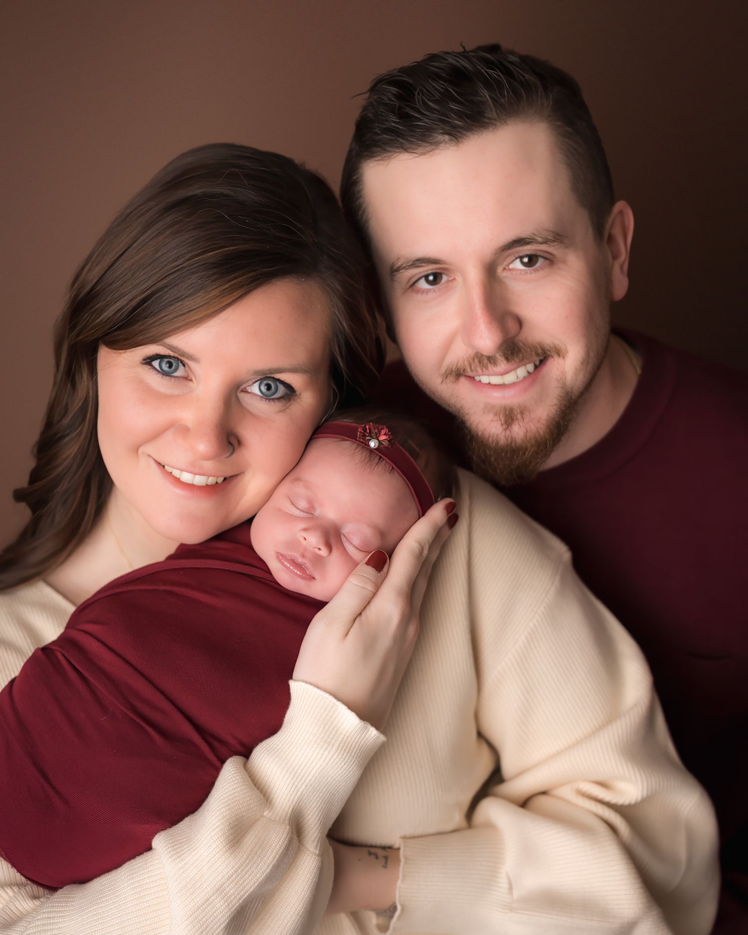Smiling couple holding their sleeping newborn baby wrapped in a cream and red blanket during a studio portrait session.