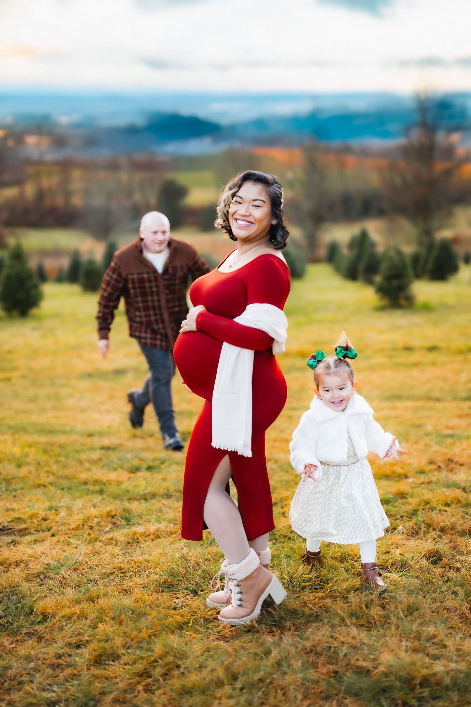 Pregnant woman in red dress poses with toddler girl and husband at Christmas tree farm during holiday maternity photo session.