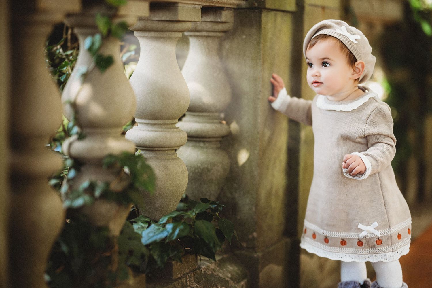 Cute toddler girl in beige knit dress and beret touching stone balustrade in an elegant garden setting.