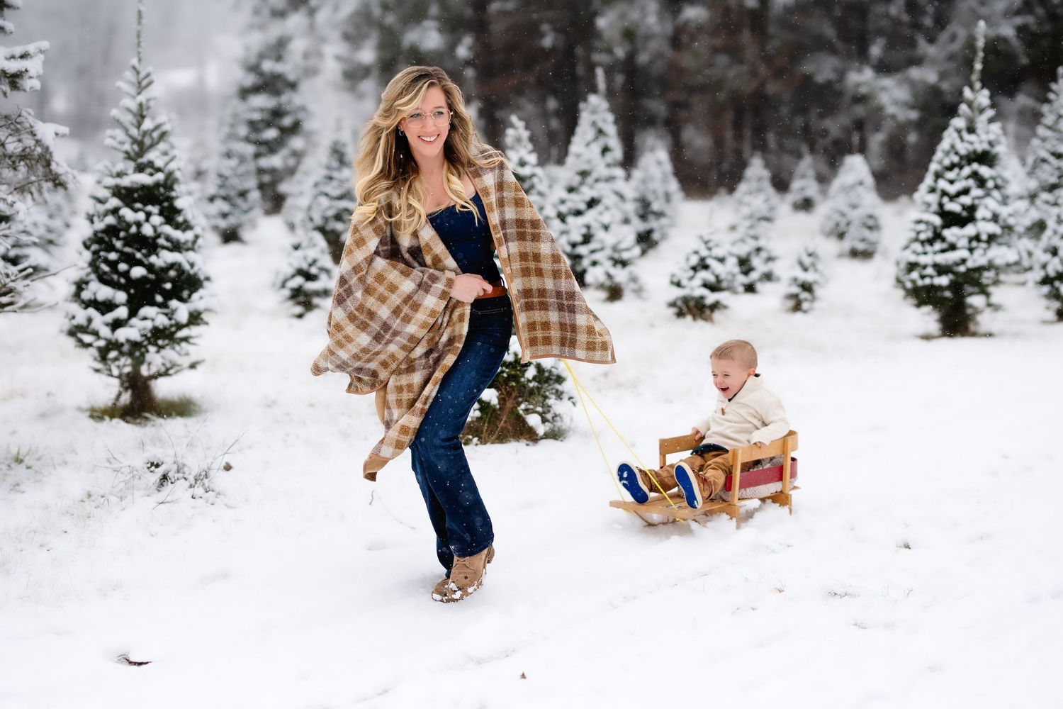 Joyful mother in plaid blanket wrap pulling laughing toddler on wooden sled through snowy Christmas tree farm.