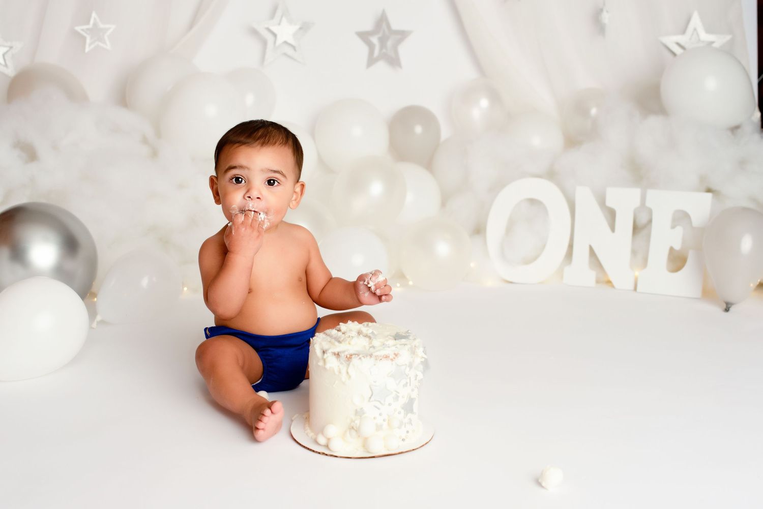 Baby boy celebrates first birthday with white smash cake, balloons, and ONE sign in bright studio setting.