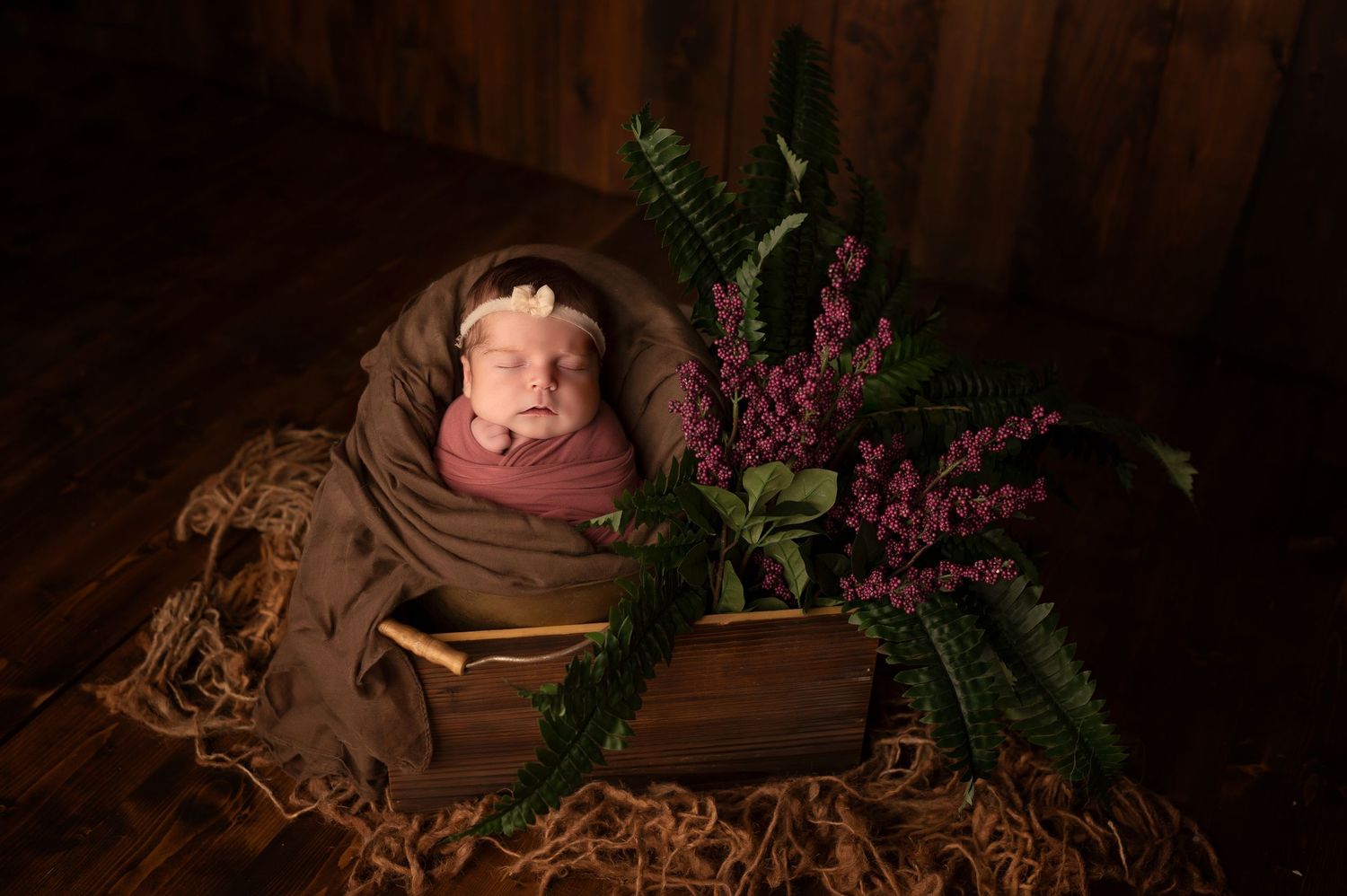 Newborn baby wrapped in brown swaddle sleeping in wooden box with ferns and purple flowers.