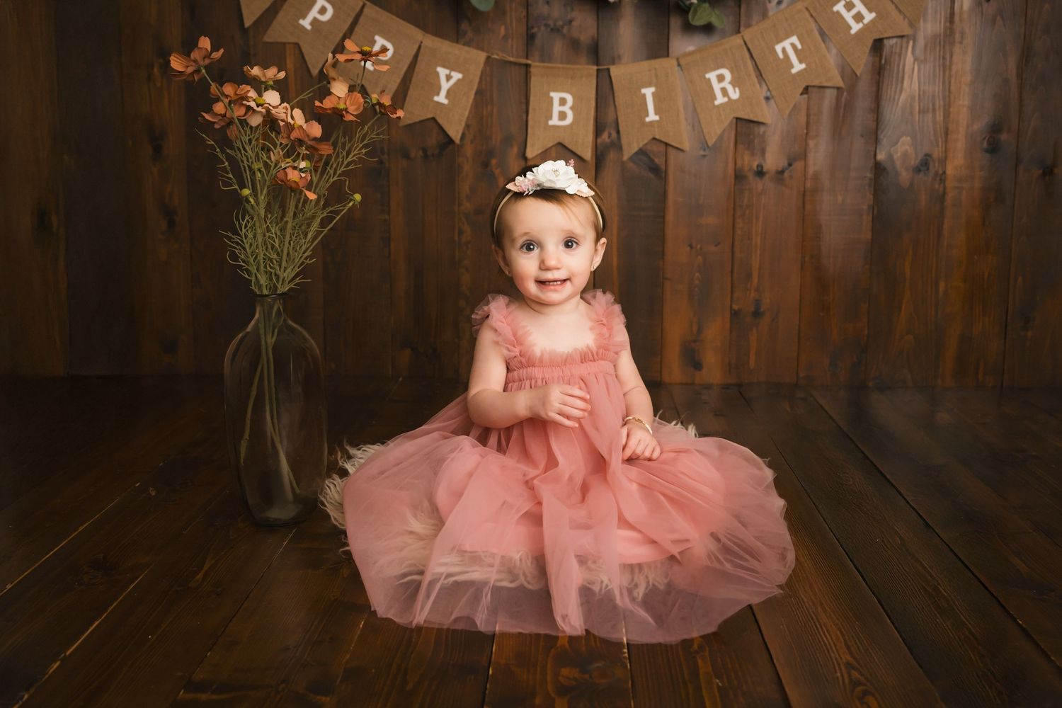 Baby girl in pink tutu dress sits on wood floor before Happy Birthday banner for first birthday portrait.