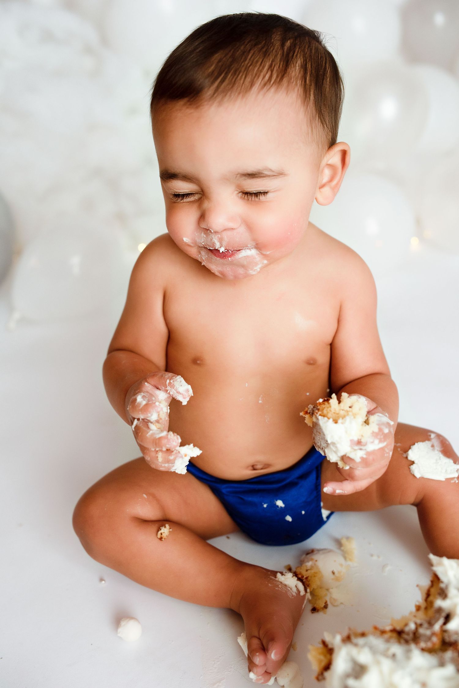 Baby boy in blue diaper smashing first birthday cake, covered in frosting with eyes closed in delight.