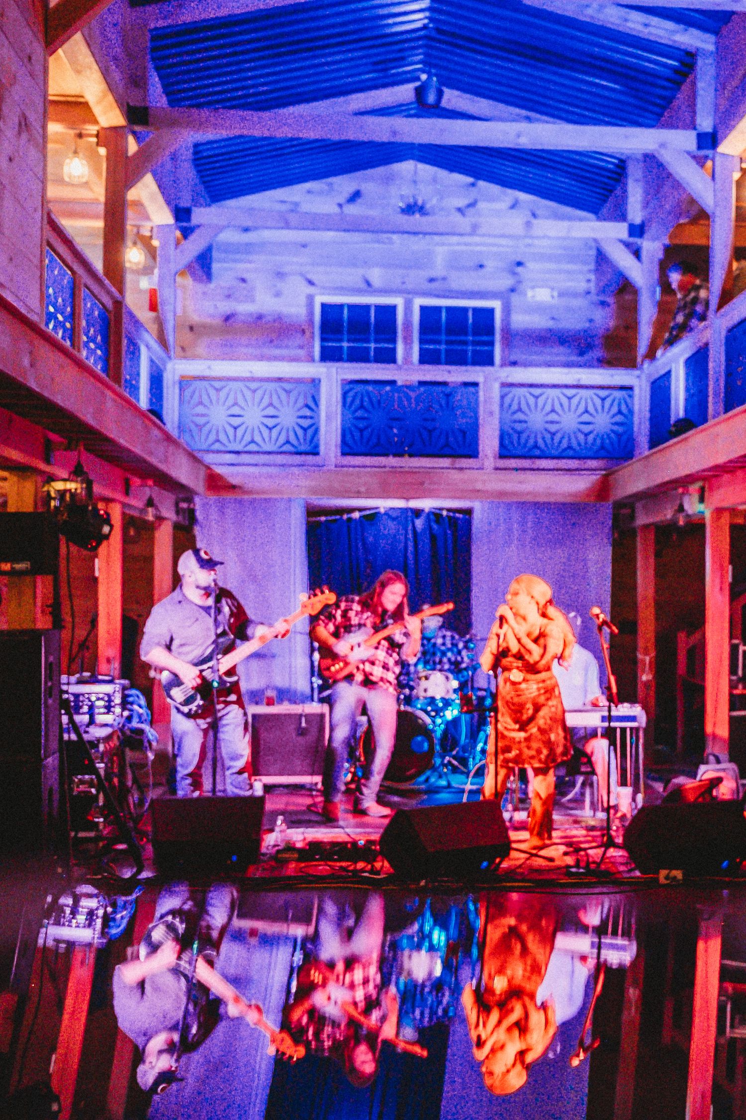 Live band performing on a rustic indoor stage with vibrant purple and blue lighting reflecting off a glossy floor.