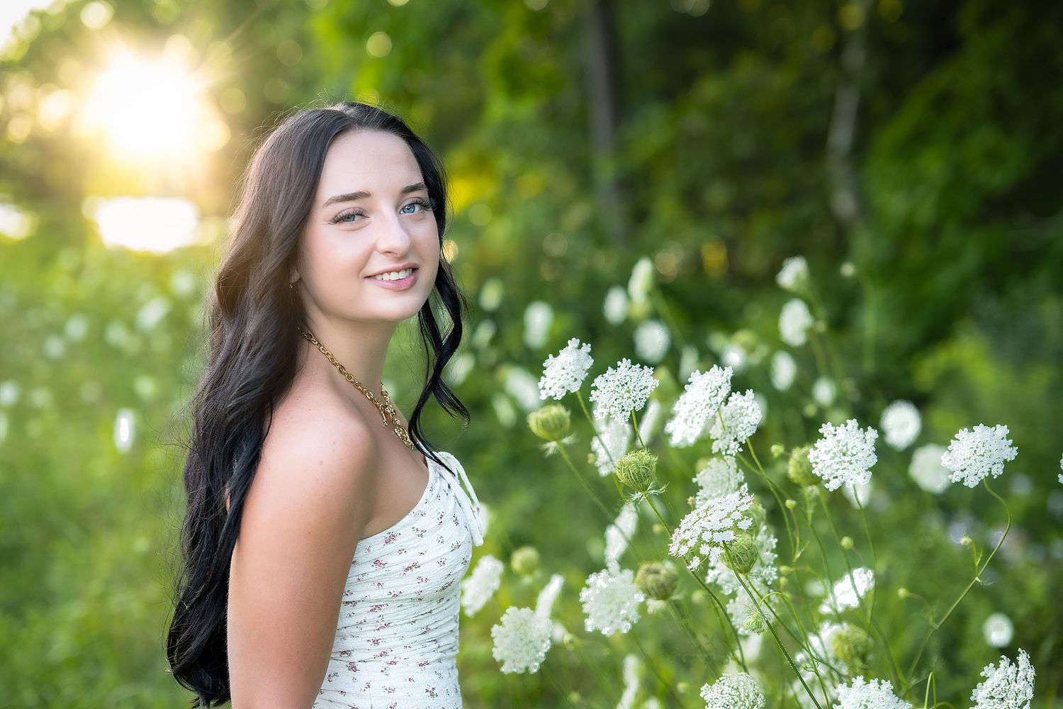 Young woman in white dress smiling among white wildflowers in a sunlit green garden.