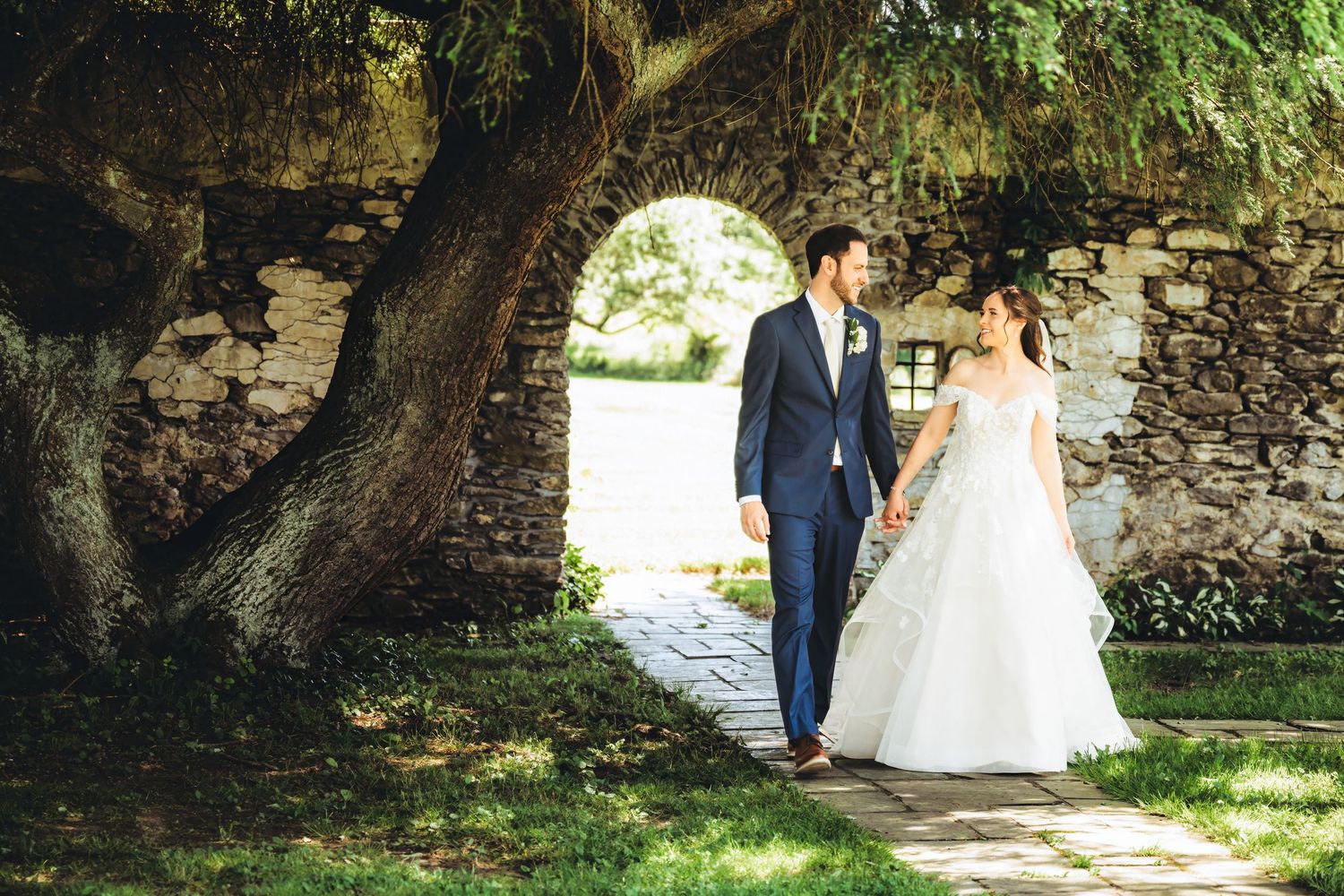 Bride and groom hold hands walking under large tree by stone arch wall on sunny wedding day.
