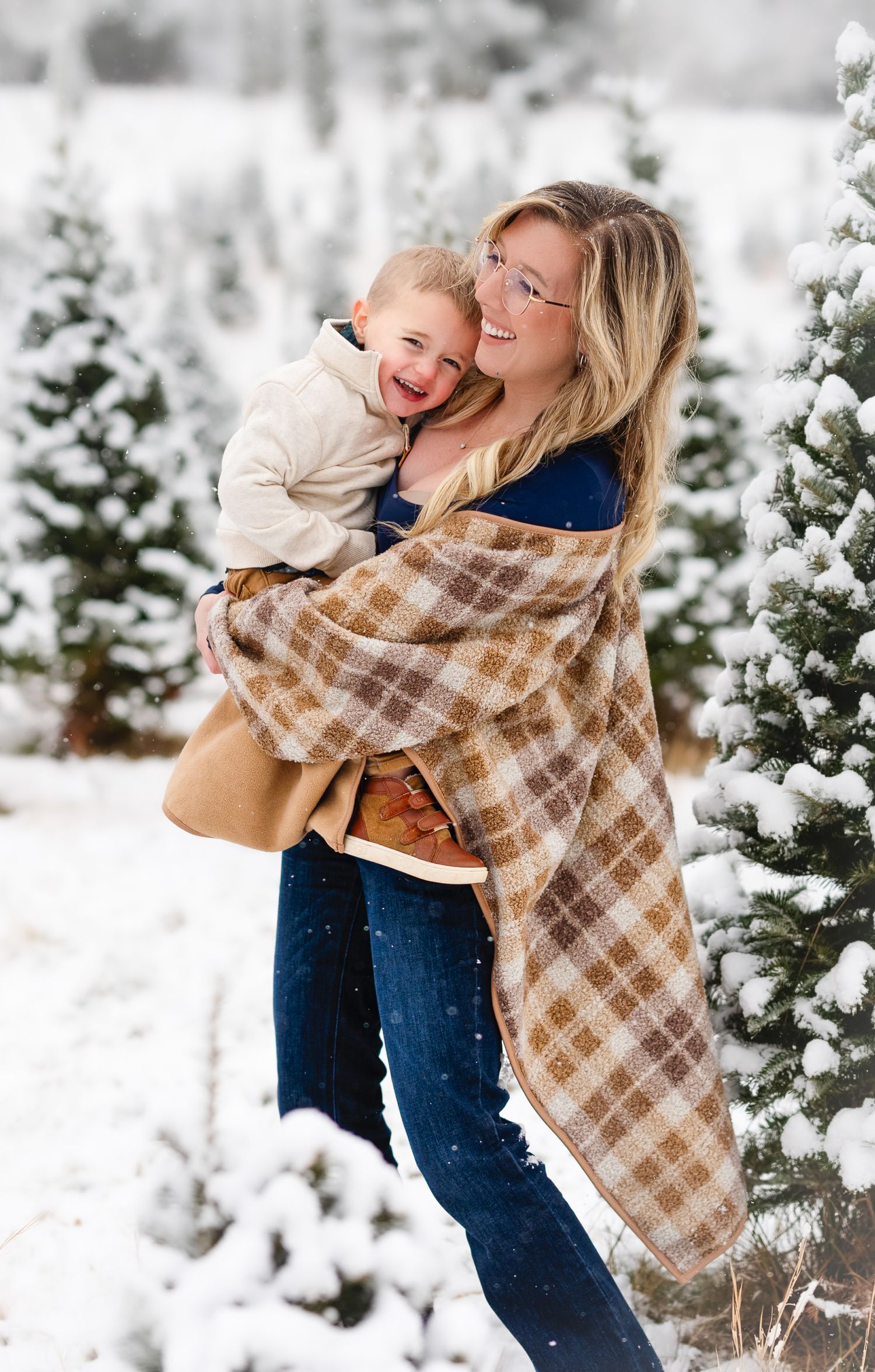 Woman wrapped in plaid blanket scarf holds laughing toddler at snowy Christmas tree farm.