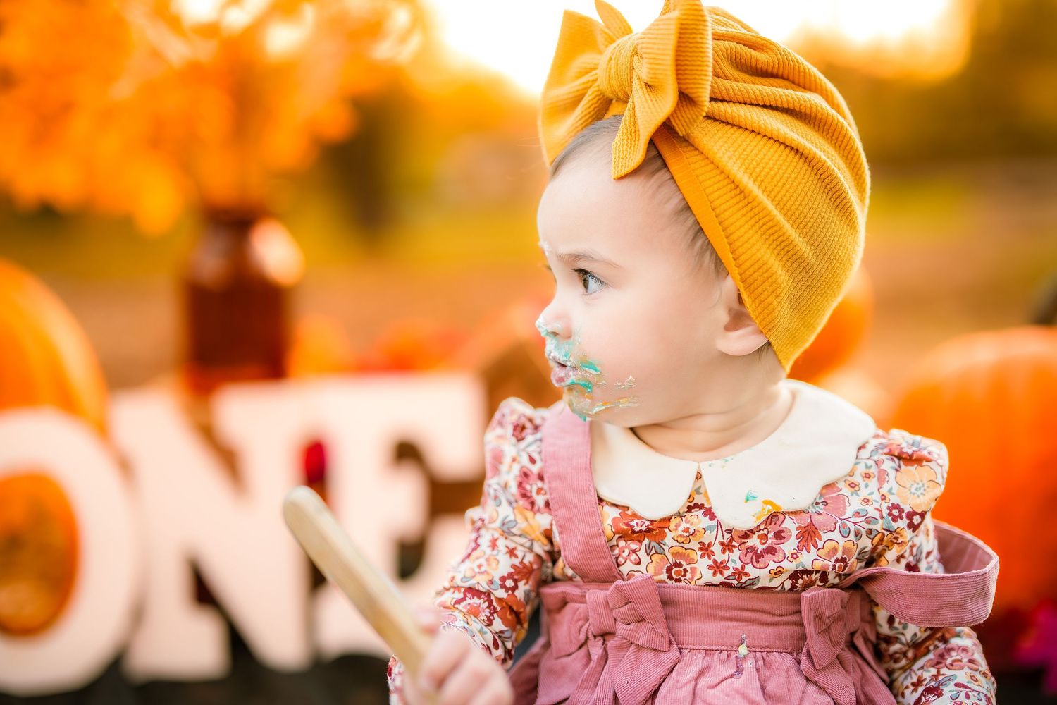Adorable baby girl in mustard yellow turban bow and floral dress at autumn fall pumpkin first birthday photo session.