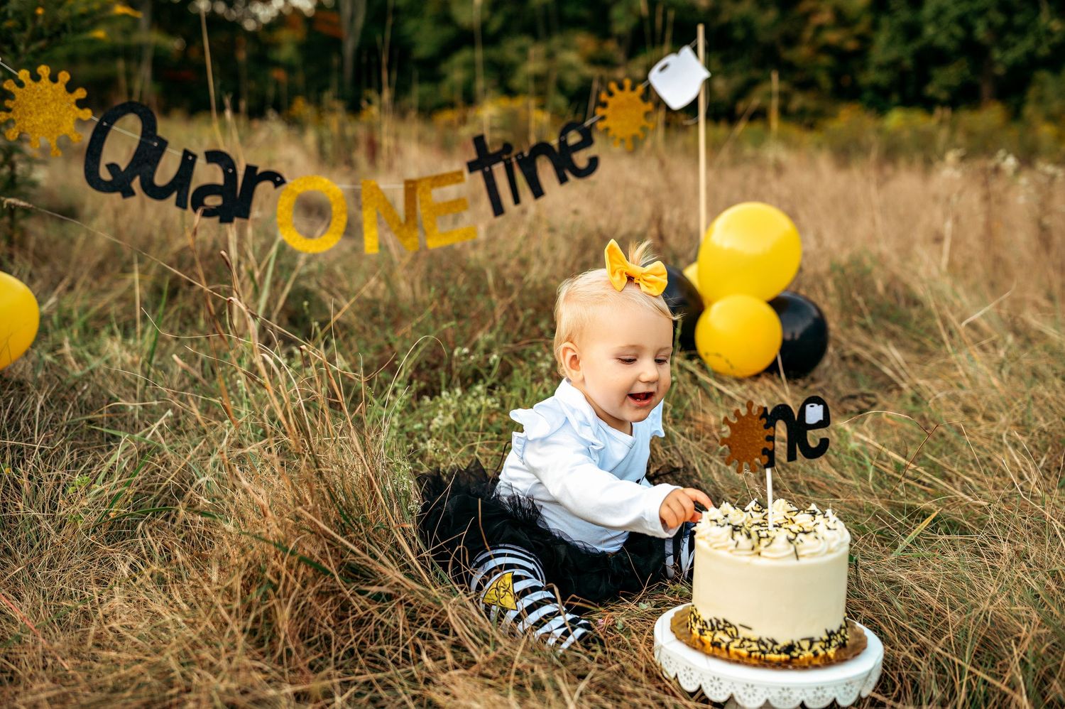 Baby's first birthday cake smash outdoors with QuarantineONE banner, yellow balloons, and white cake in a field.