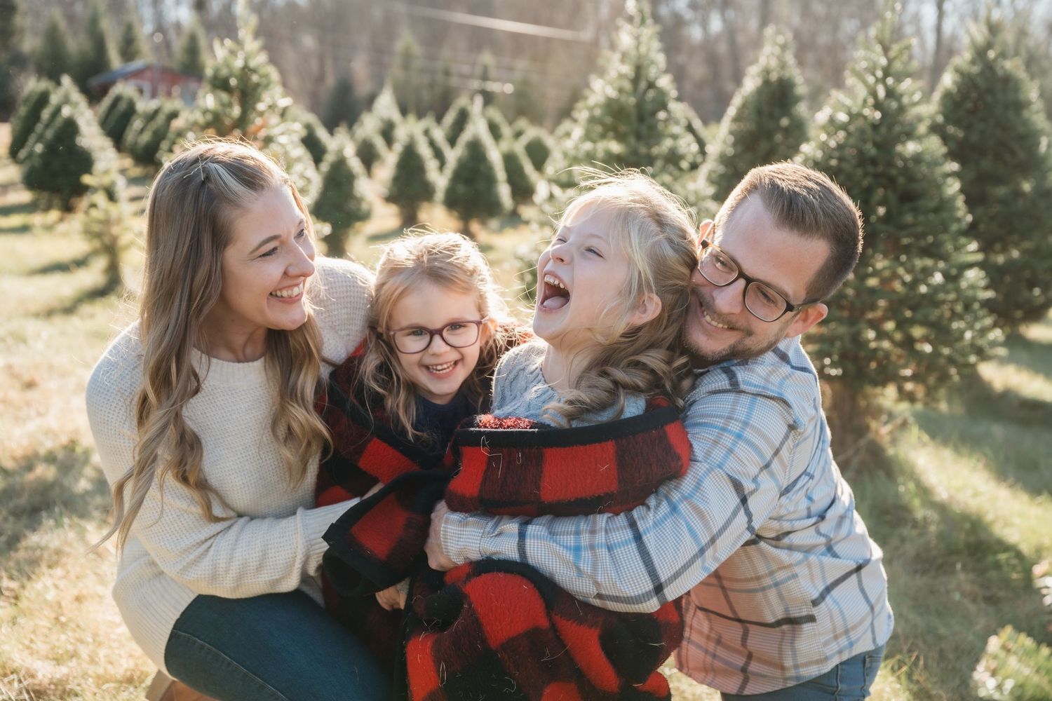 Happy family of four laughing together at a Christmas tree farm on a sunny winter day.