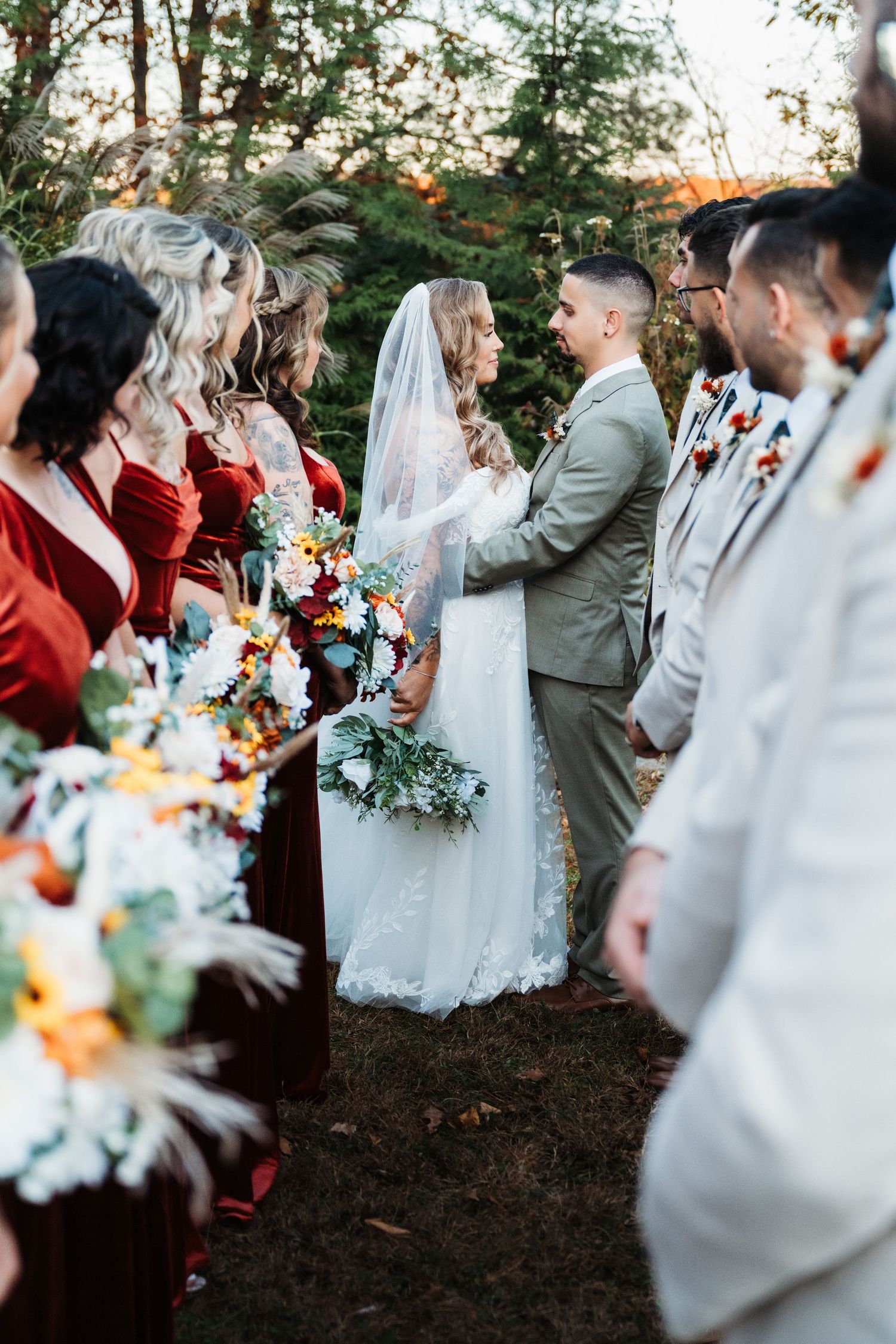 Bride and groom exchange vows outdoors flanked by bridesmaids in red velvet gowns and groomsmen in tan suits.