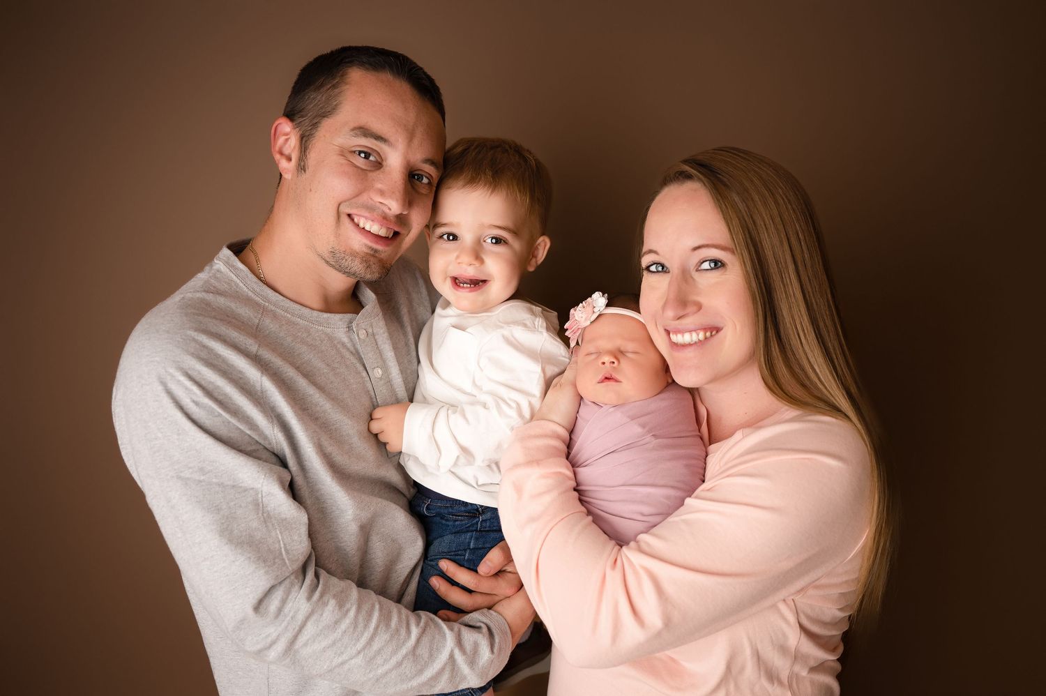 Happy family portrait with parents holding toddler and newborn baby against brown studio background.