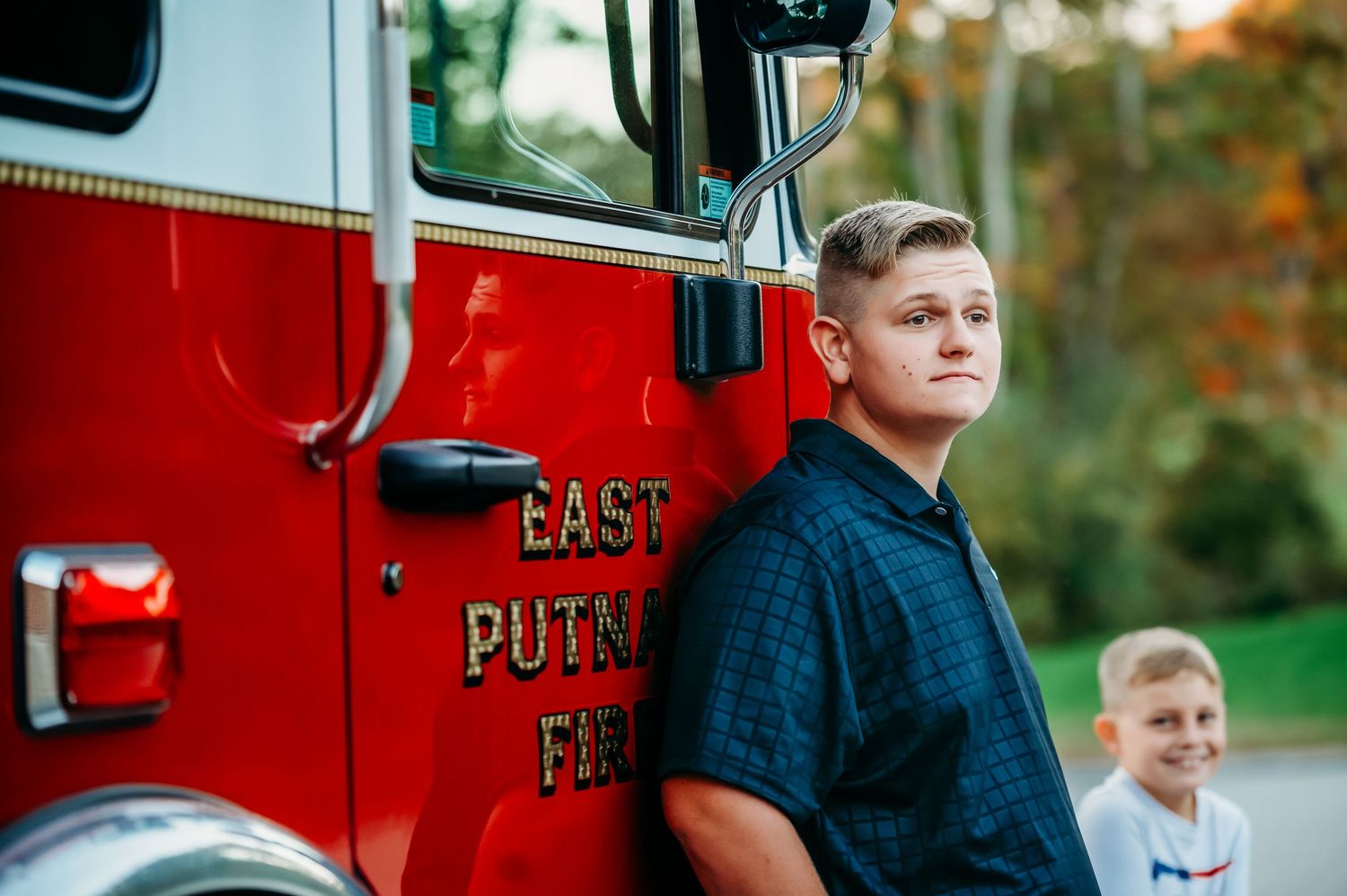 Two boys pose beside a red East Putnam fire truck outdoors, with the older teen leaning casually against it.
