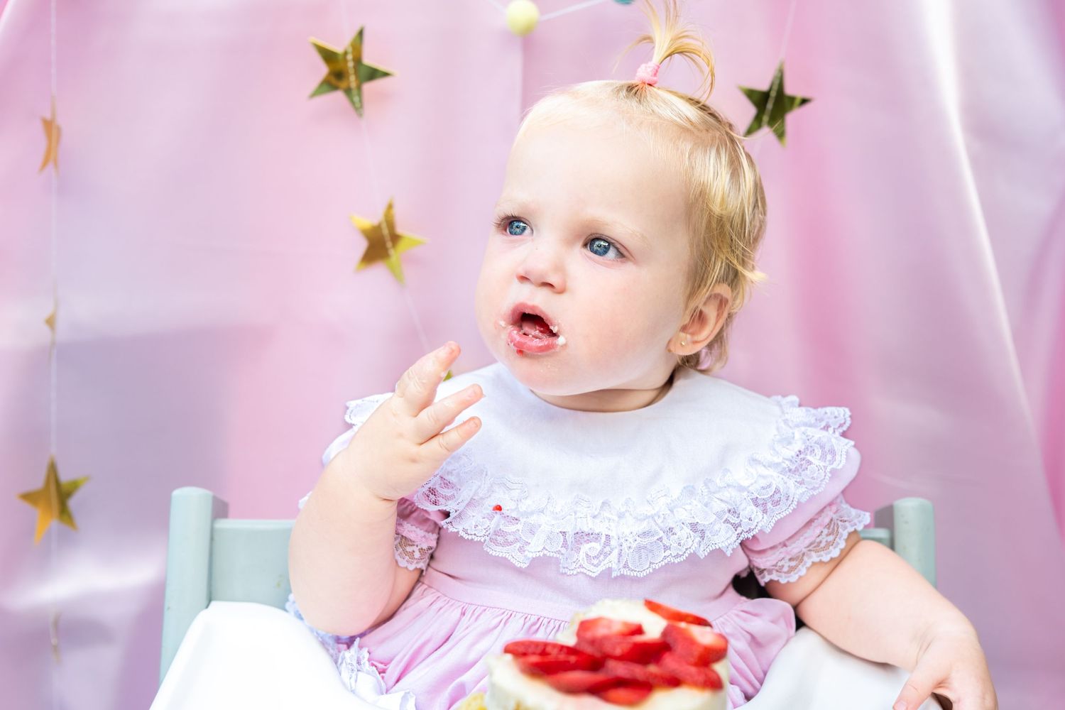 Baby girl in pink dress eating strawberries in high chair at first birthday party with gold star decorations.