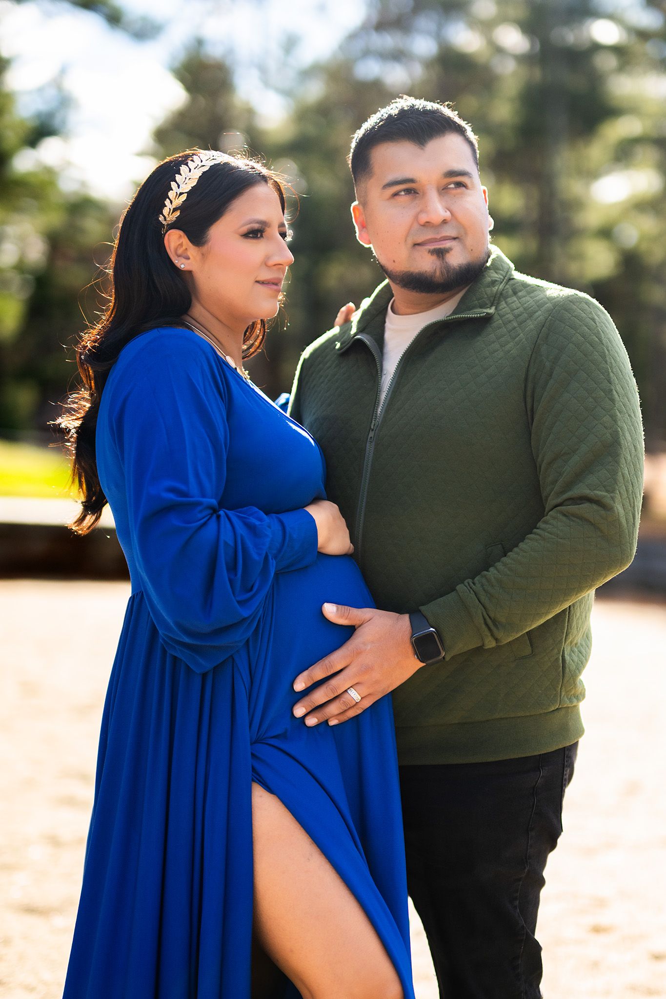 Pregnant woman in blue dress with partner in green shirt posing outdoors in sunny park setting.