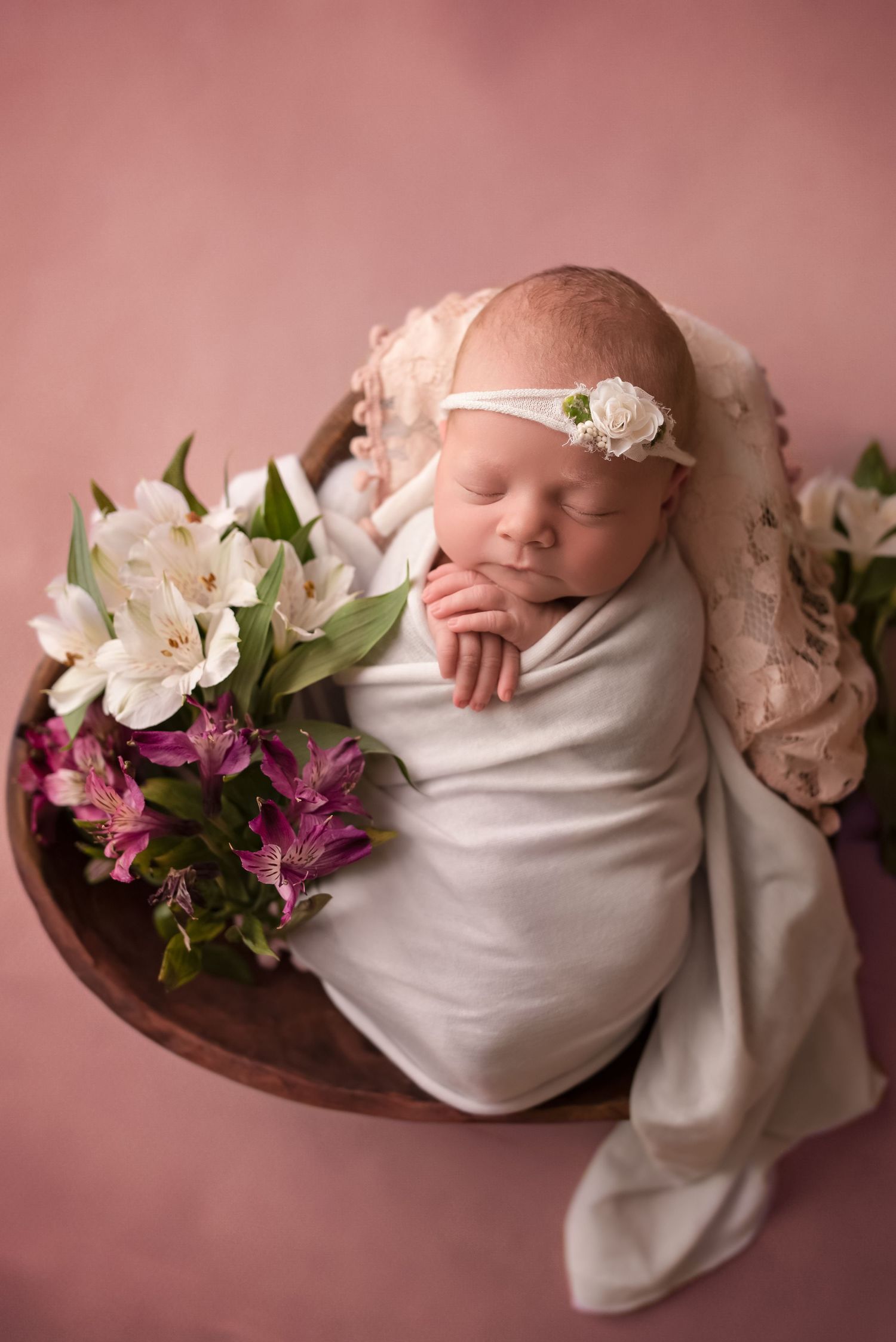 Sleeping newborn baby girl swaddled in white wrap with floral headband, nestled in wooden bowl with pink and white flowers.