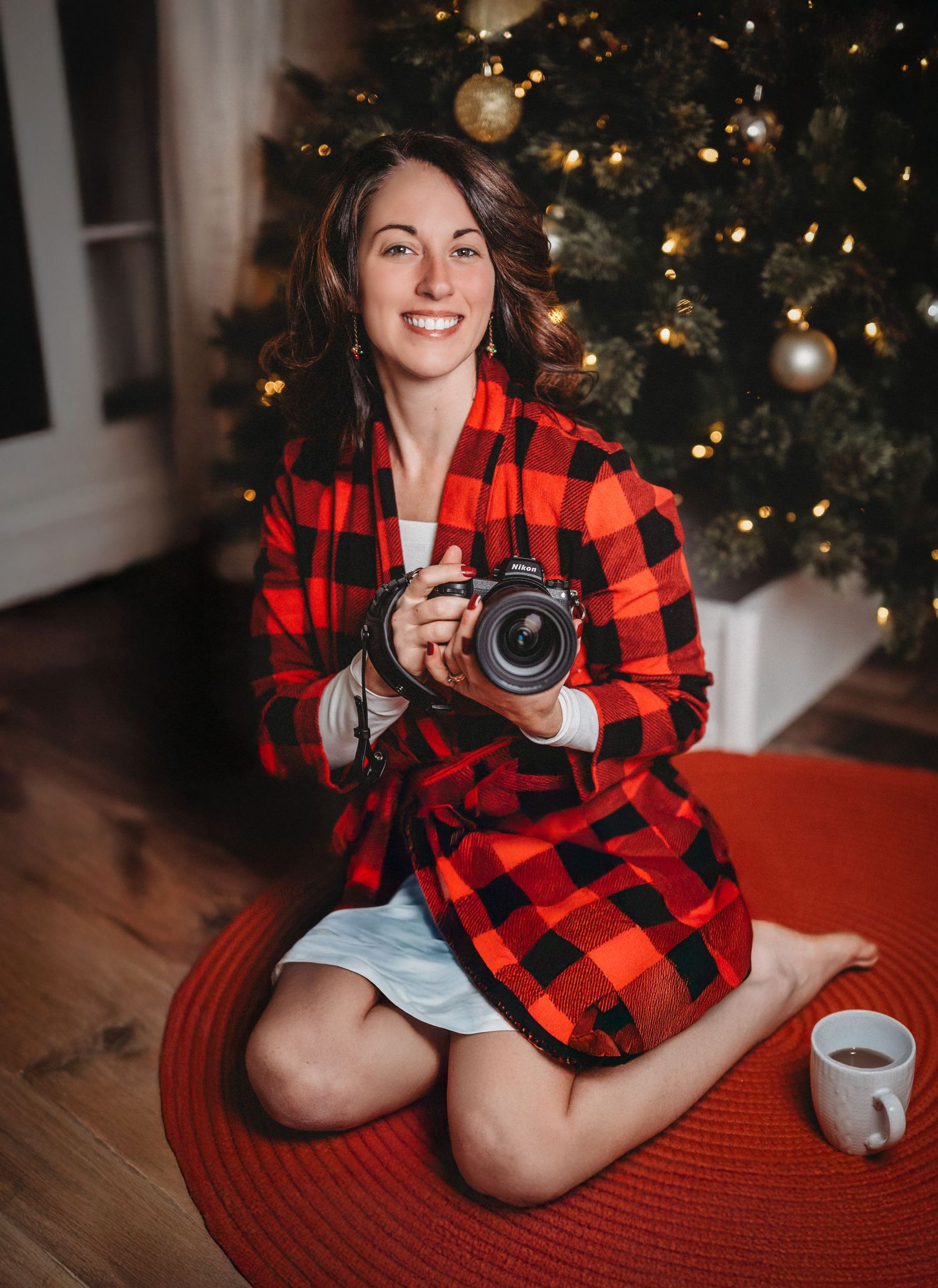 Woman in red plaid robe holds camera, sits cross-legged by decorated Christmas tree with coffee cup.