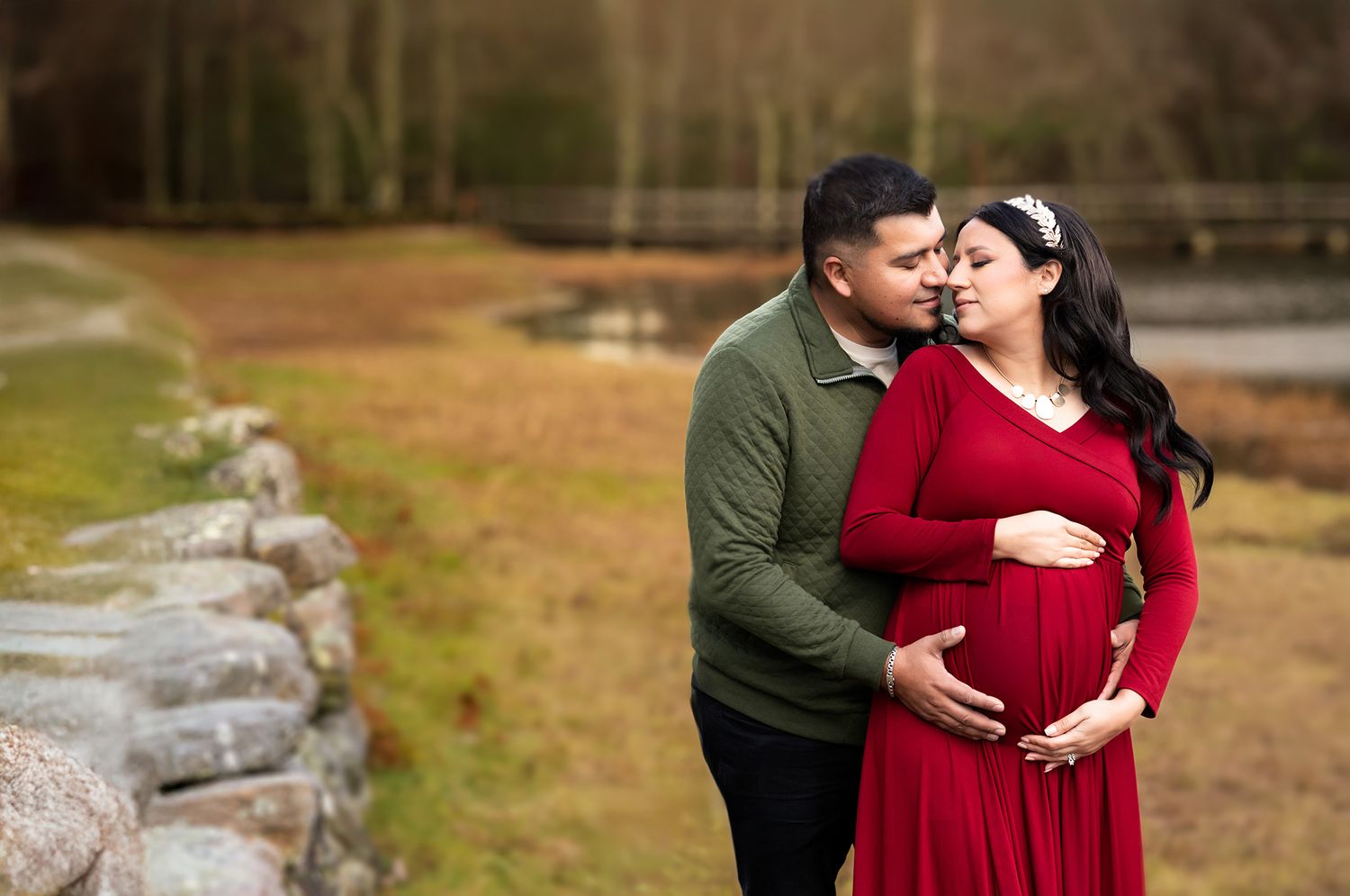 Couple sharing tender moment outdoors, pregnant woman in red dress cradling baby bump, partner in green sweater kissing her cheek.