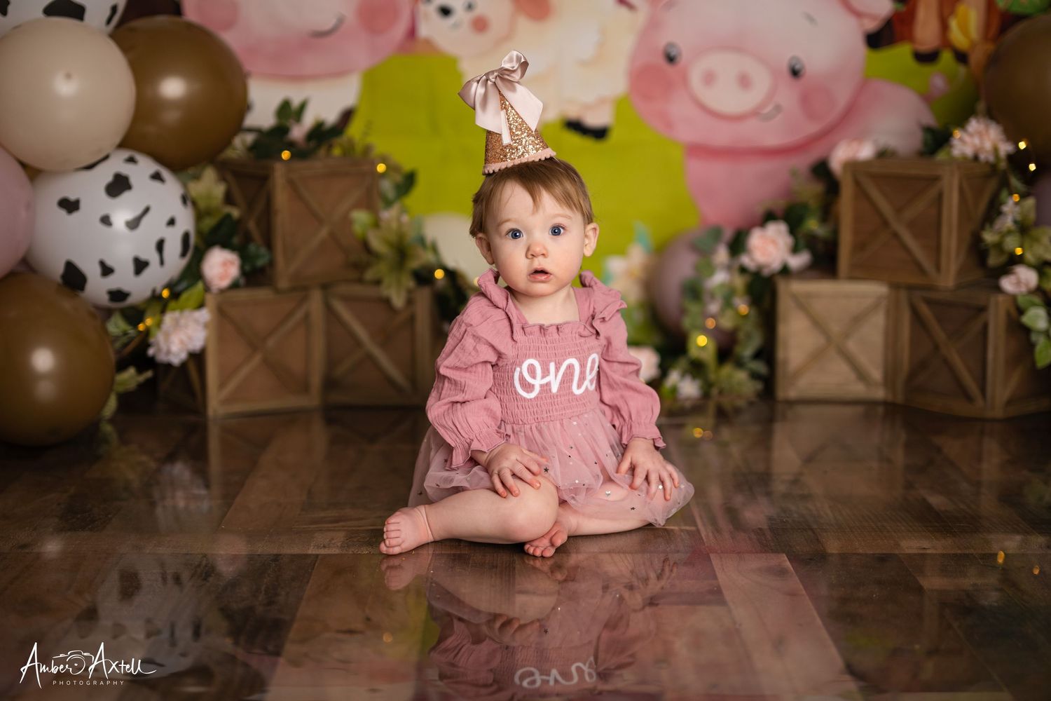 Baby girl in pink dress with party hat sits on floor at farm-themed first birthday cake smash photo session.