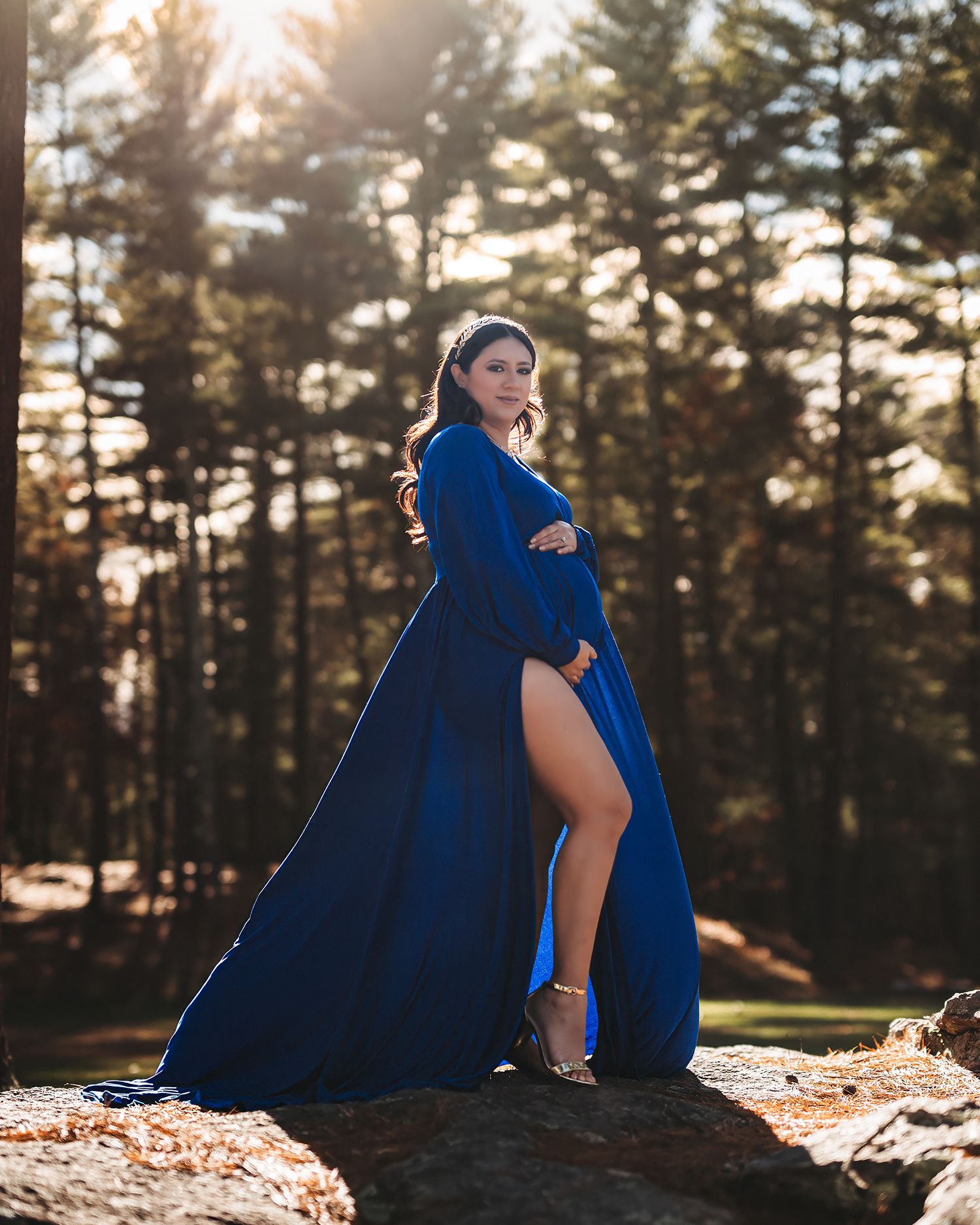 Pregnant woman in flowing royal blue maternity gown posing on rocks in sunlit forest.