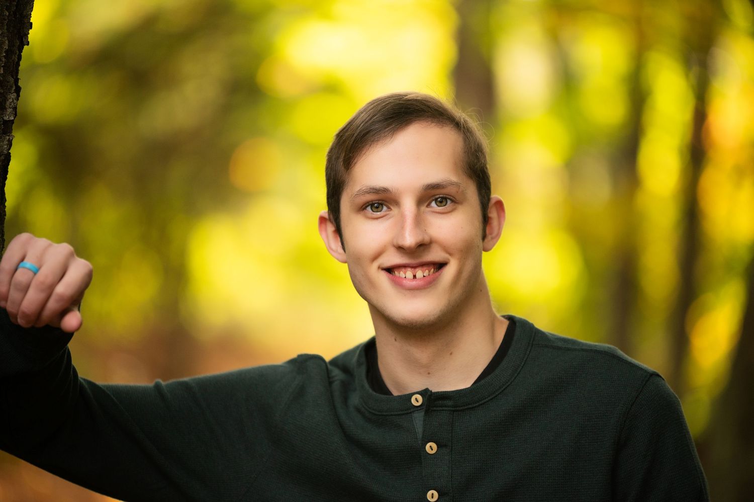 Smiling young man in dark green shirt posing outdoors among vibrant yellow-green autumn foliage.