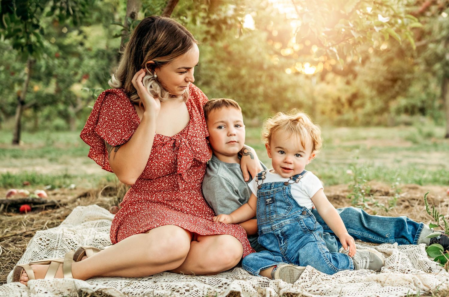 A mother in a red floral dress sits outdoors with her two young children on a blanket in a sunlit park.