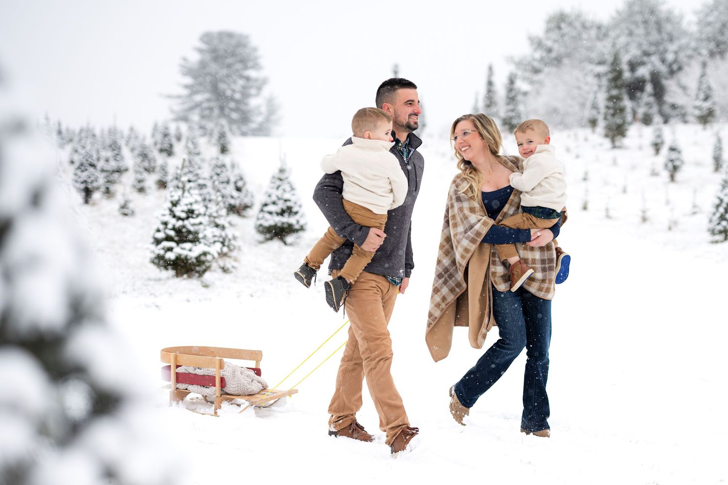 Family walking through snowy Christmas tree farm, parents carrying toddlers with a wooden sled in a winter wonderland.